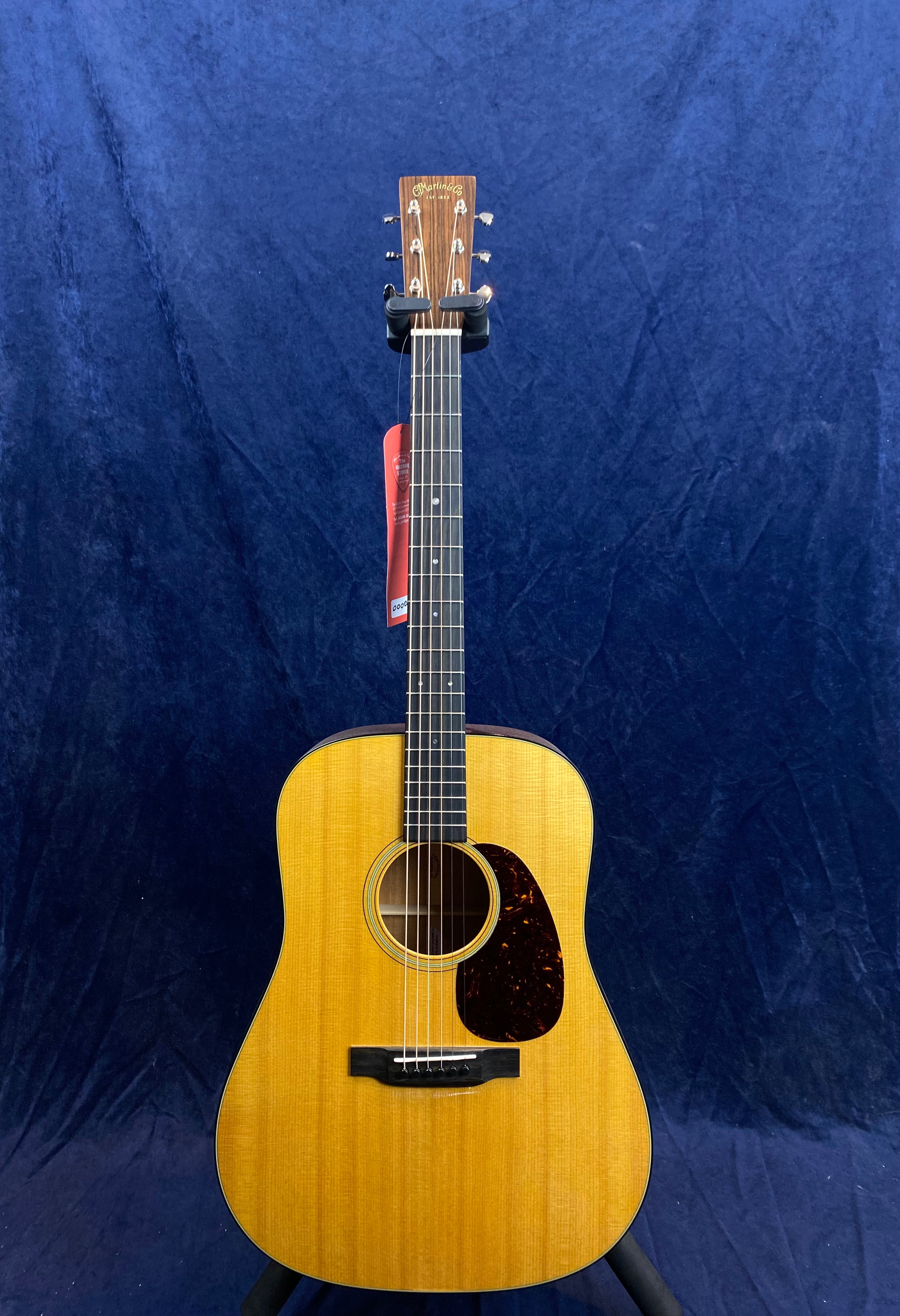Acoustic guitar with a light wood body, black fretboard, six strings, tortoiseshell pickguard, and a red tag hanging from the headstock.