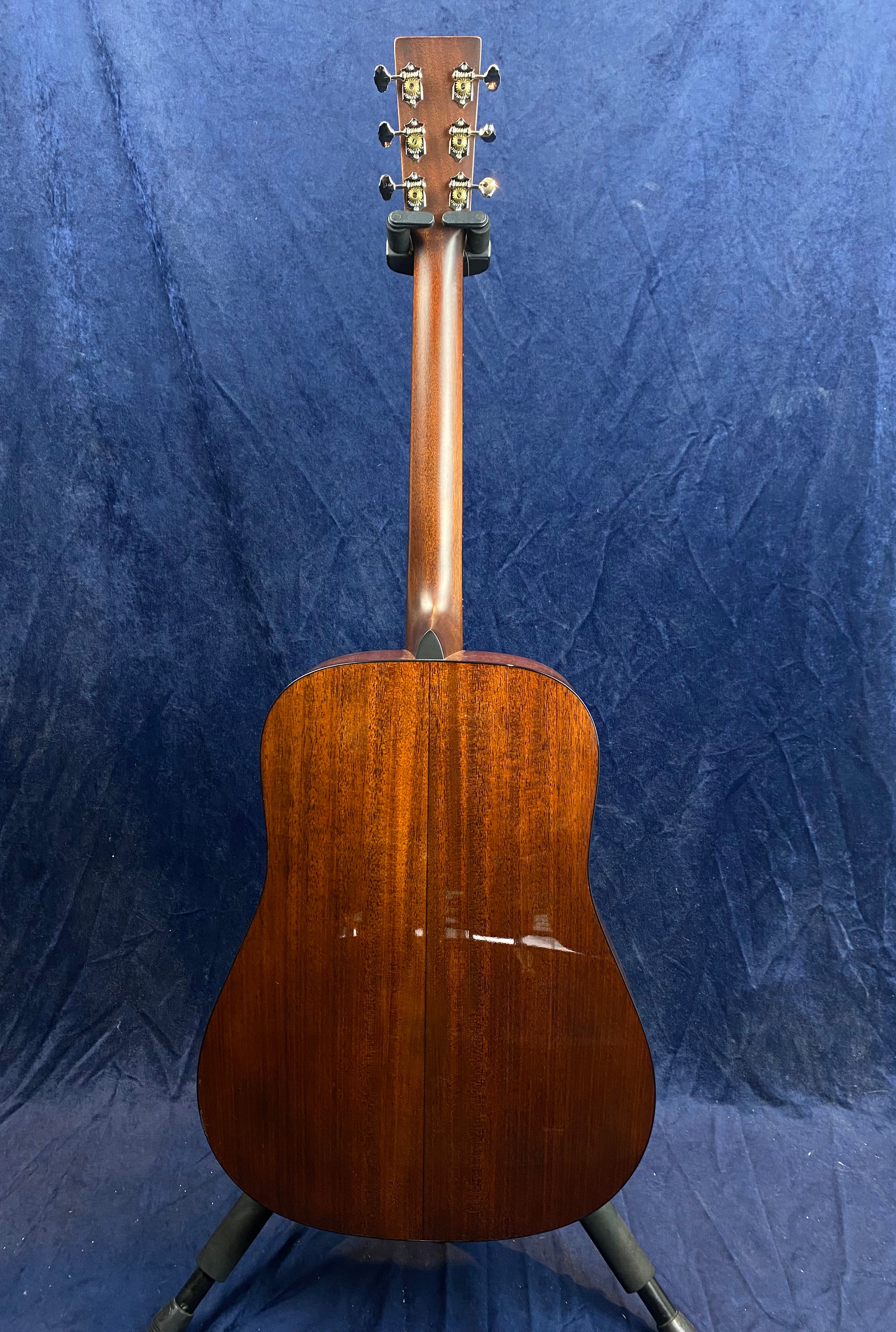 Brown acoustic guitar shown from the back, glossy wooden body with visible central seam, six tuning pegs, positioned upright on a stand.