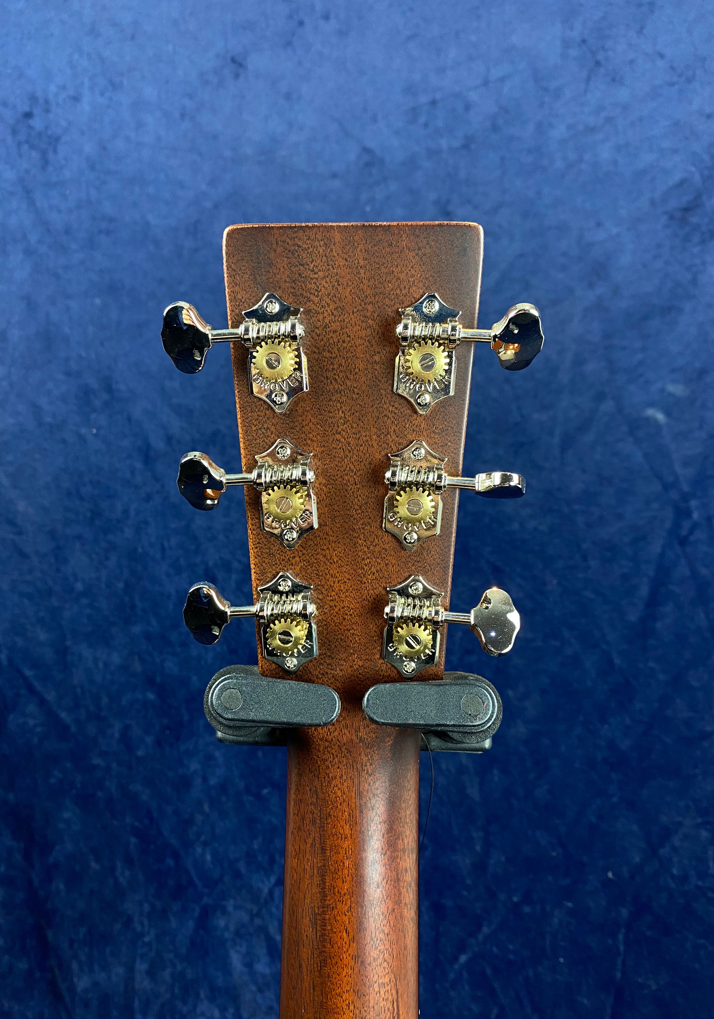 Six vintage-style metal tuning pegs are mounted on the brown wooden headstock of an acoustic guitar against a dark blue background.
