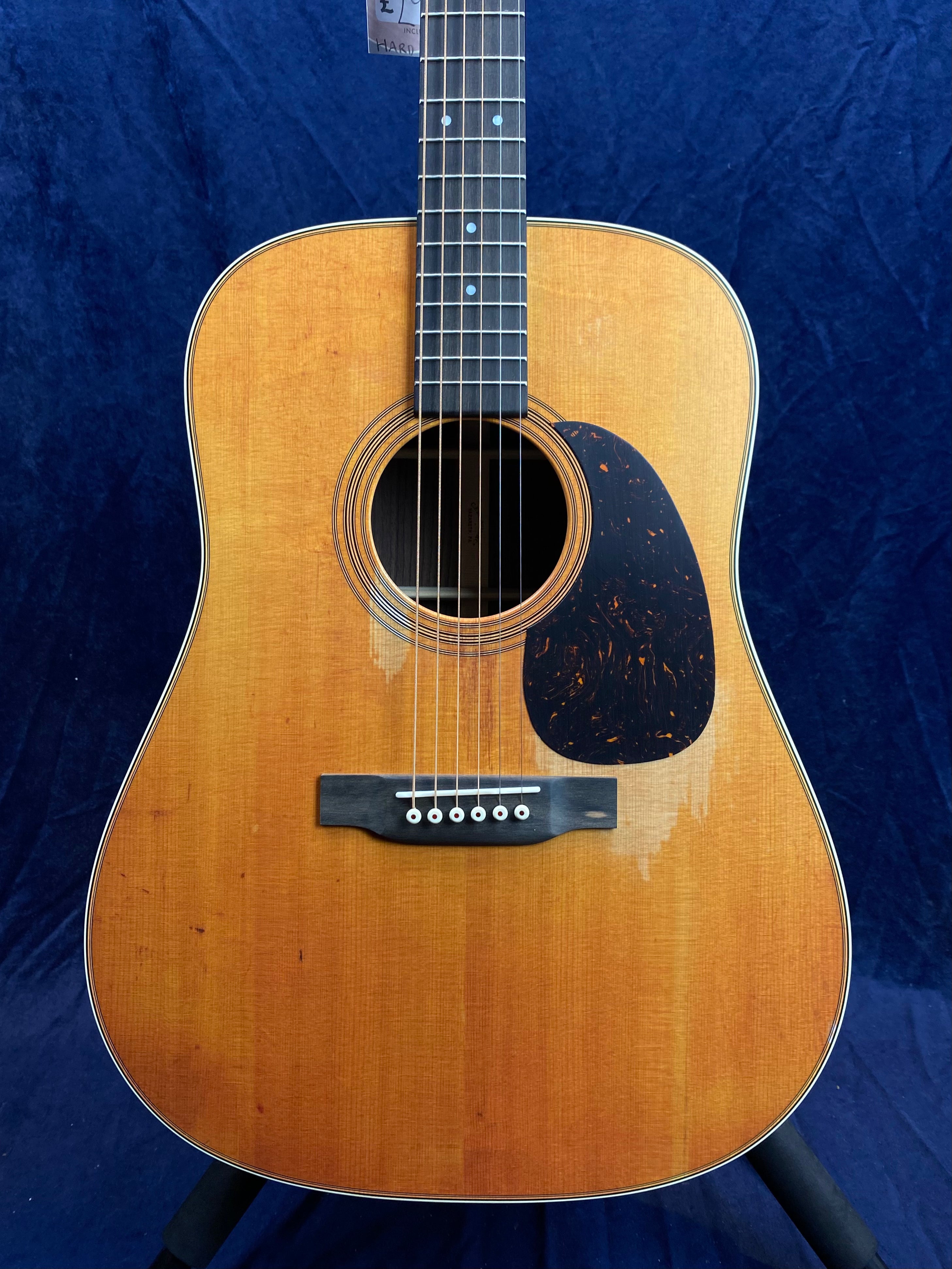 Acoustic guitar with a worn, natural wood finish and a large dark pickguard beside the soundhole, six strings, and white bridge pins.
