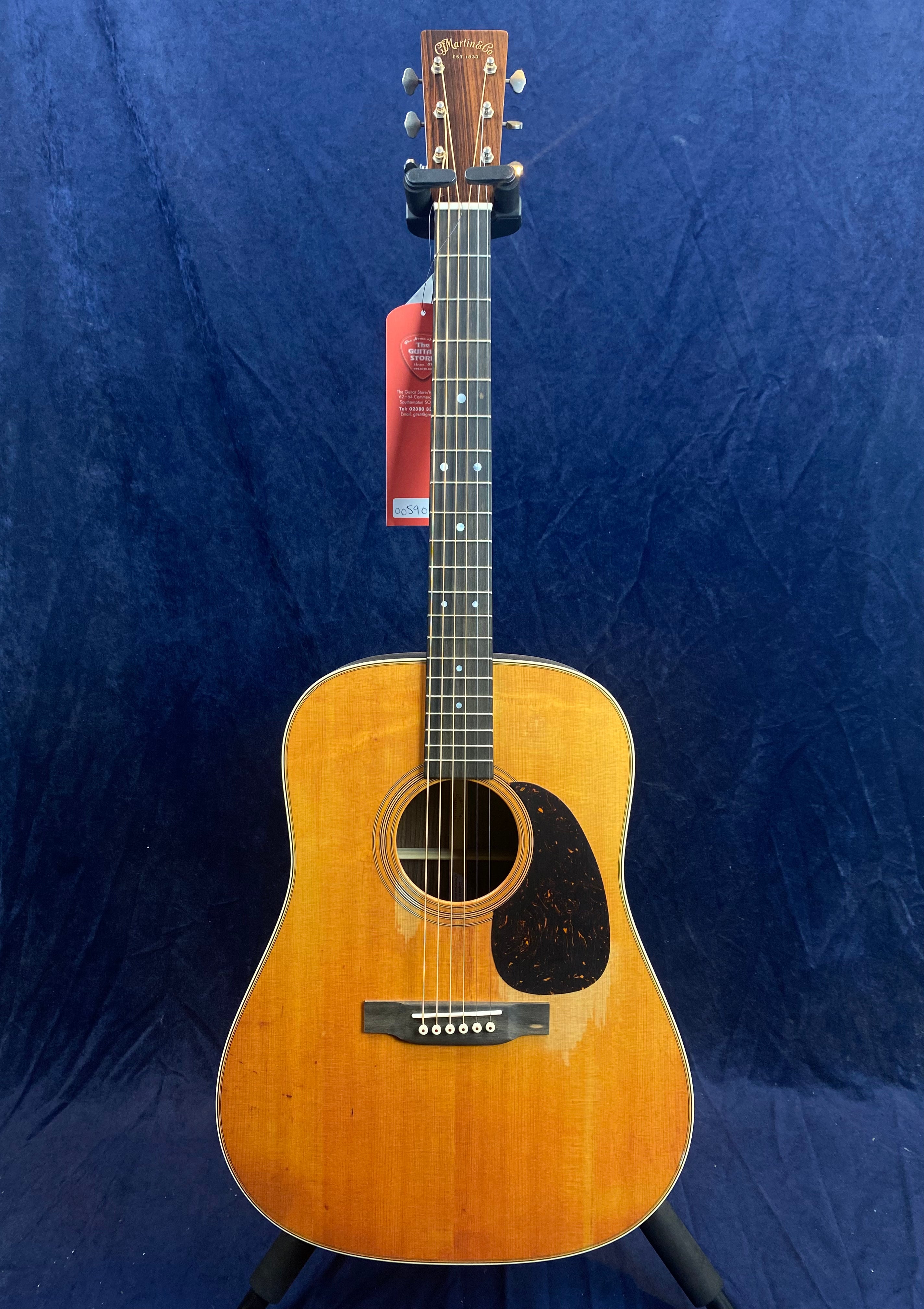 A brown acoustic guitar with a dark pickguard on the right side of the body, six strings, dot inlays on the fretboard, and a tag hanging from the headstock.
