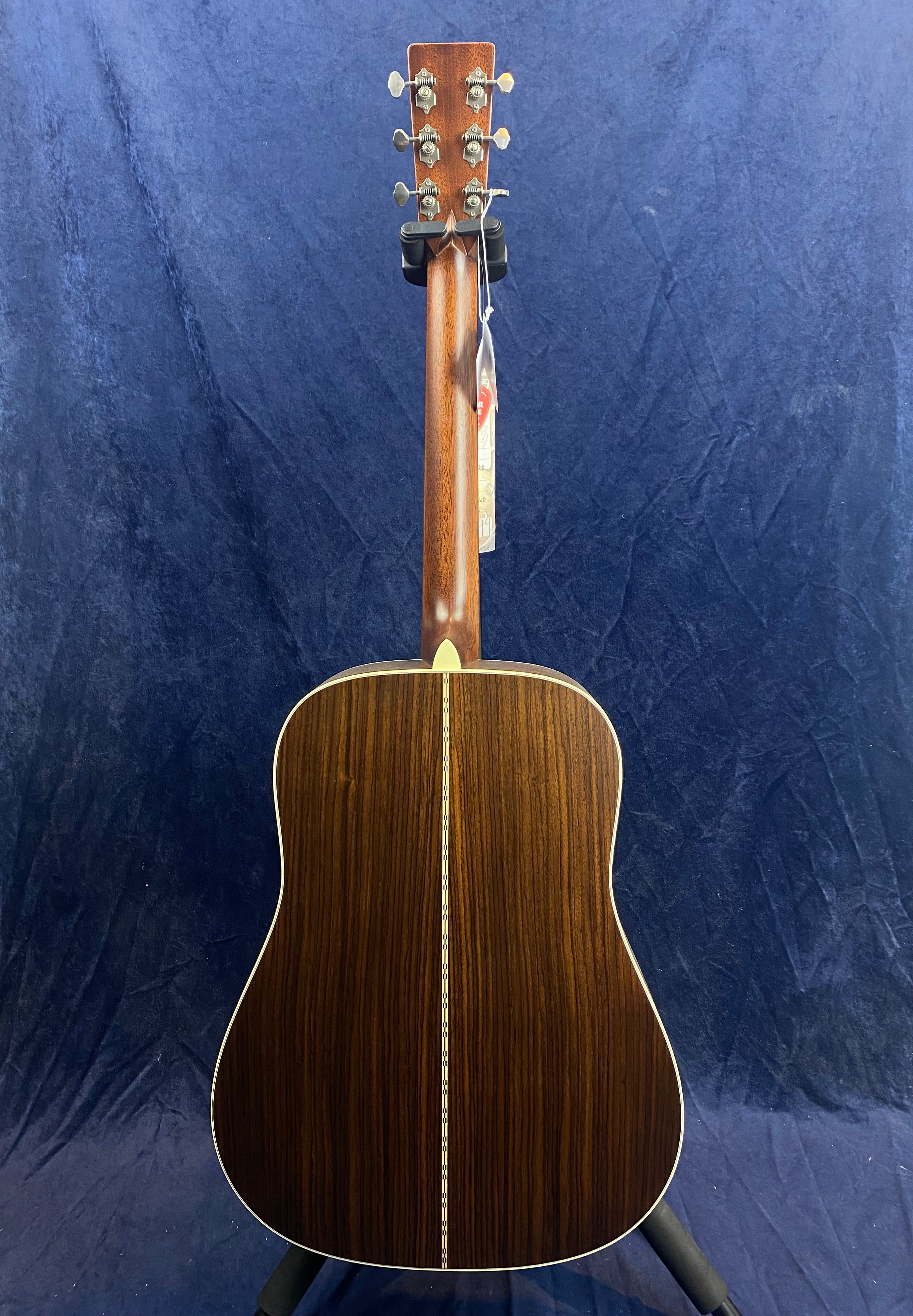 Acoustic guitar viewed from the back, showing dark wood with a decorative center strip and light binding along the edges, standing upright on a stand.