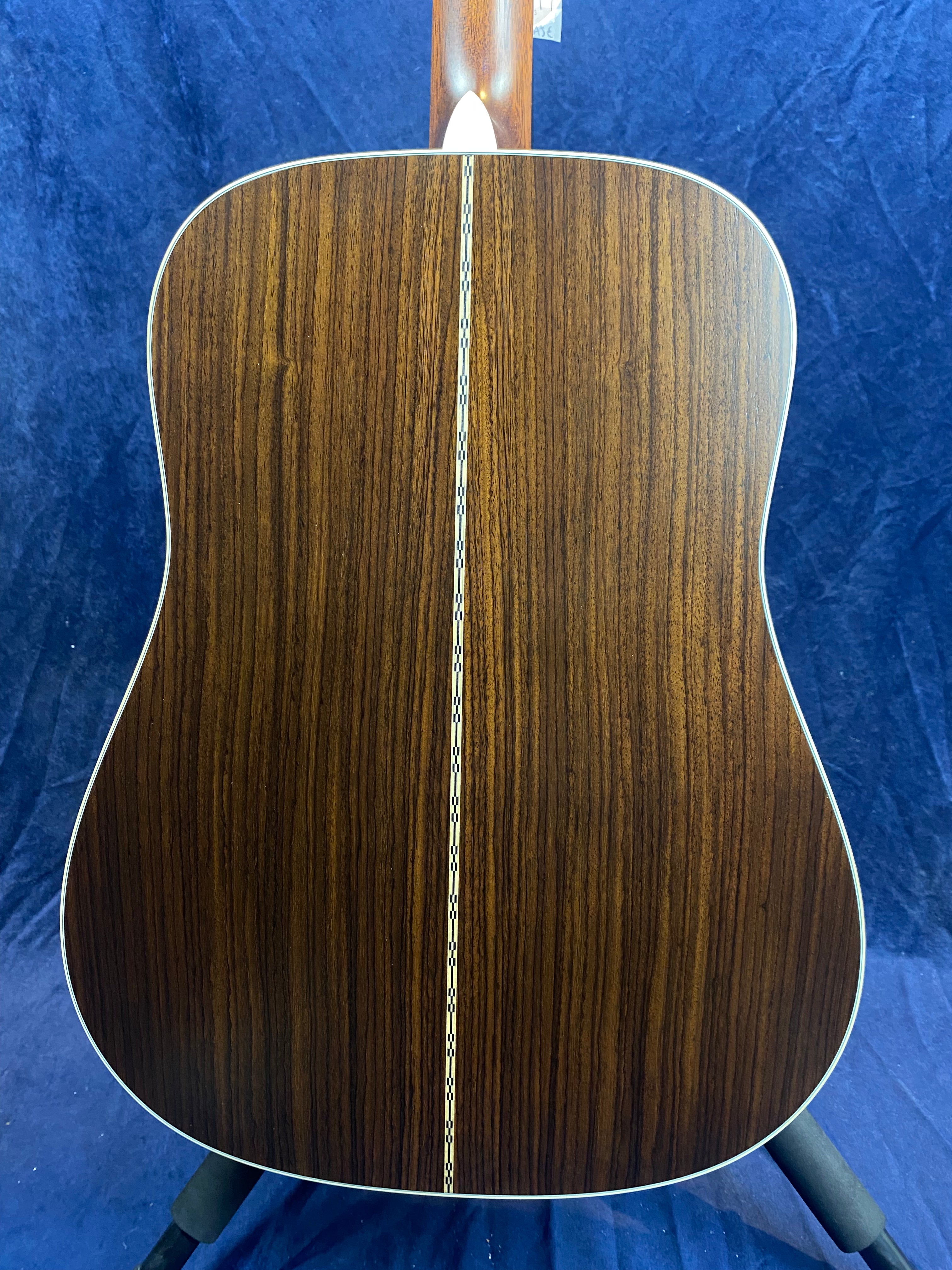 Back of an acoustic guitar with dark wood grain, a decorative light inlay runs vertically down the center, and white trim outlines the edges.