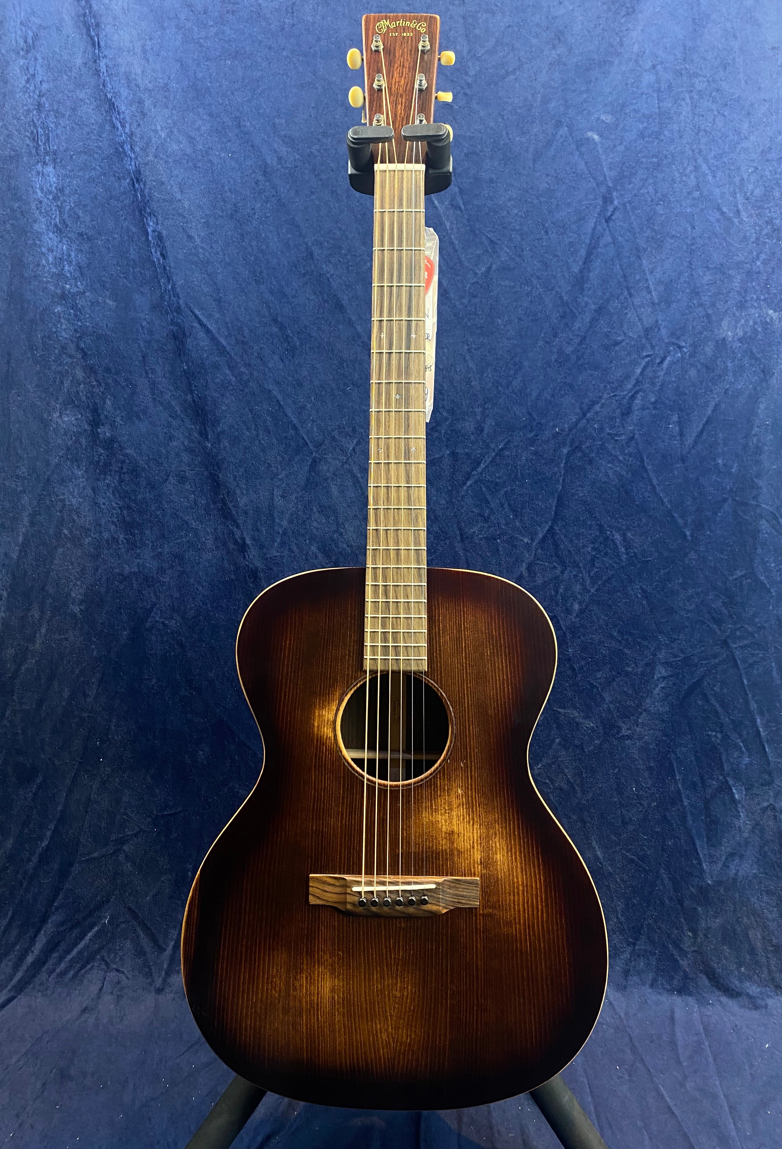 Small-bodied acoustic guitar with a dark sunburst finish, six strings, and a simple wooden bridge against a blue background.