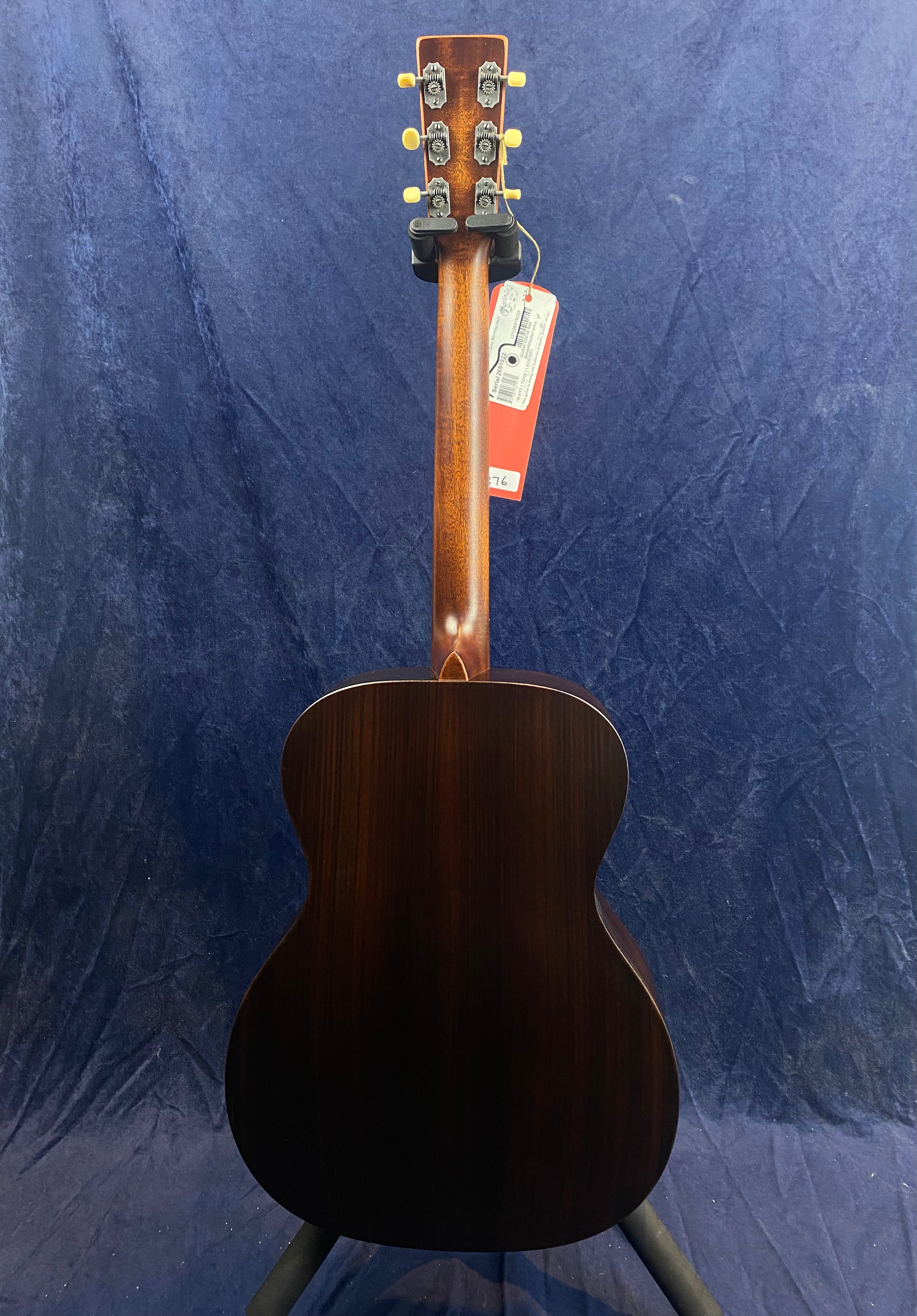 Dark wood acoustic guitar seen from the back, showing a brown neck, gold tuning pegs, and a price tag hanging near the headstock.
