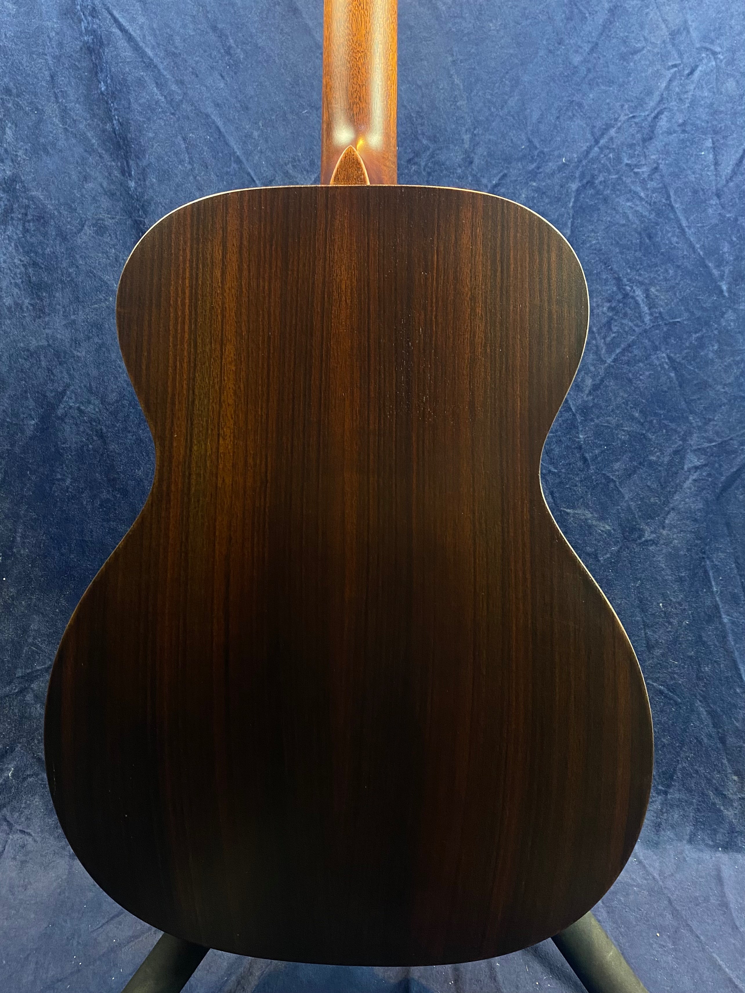 Back view of an acoustic guitar with dark wood grain, a smooth finish, and a visible neck joint against a textured dark blue background.