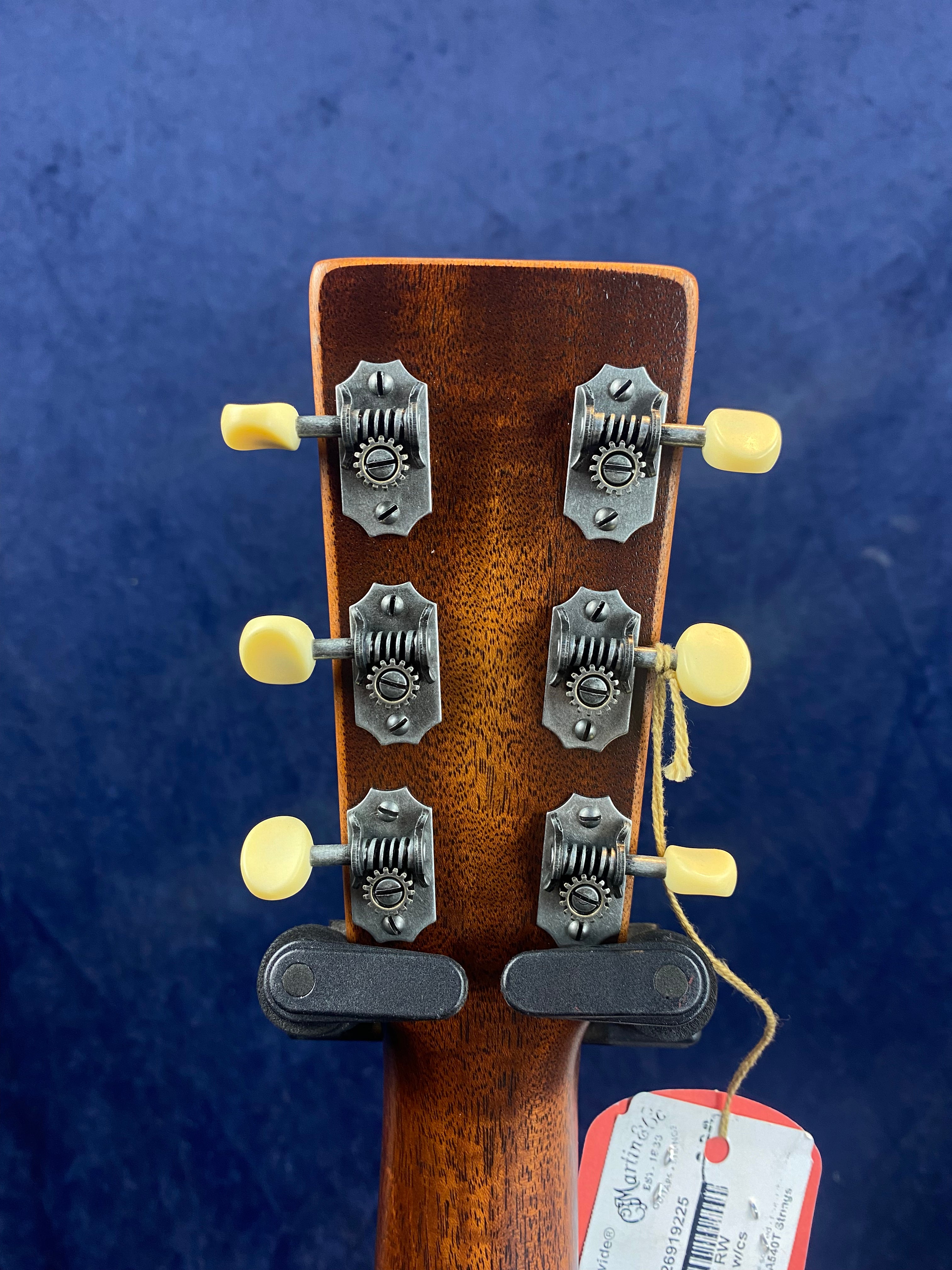 Six vintage-style tuning pegs with cream knobs on the back of a wooden acoustic guitar headstock, set against a blue background, with a hang tag attached.