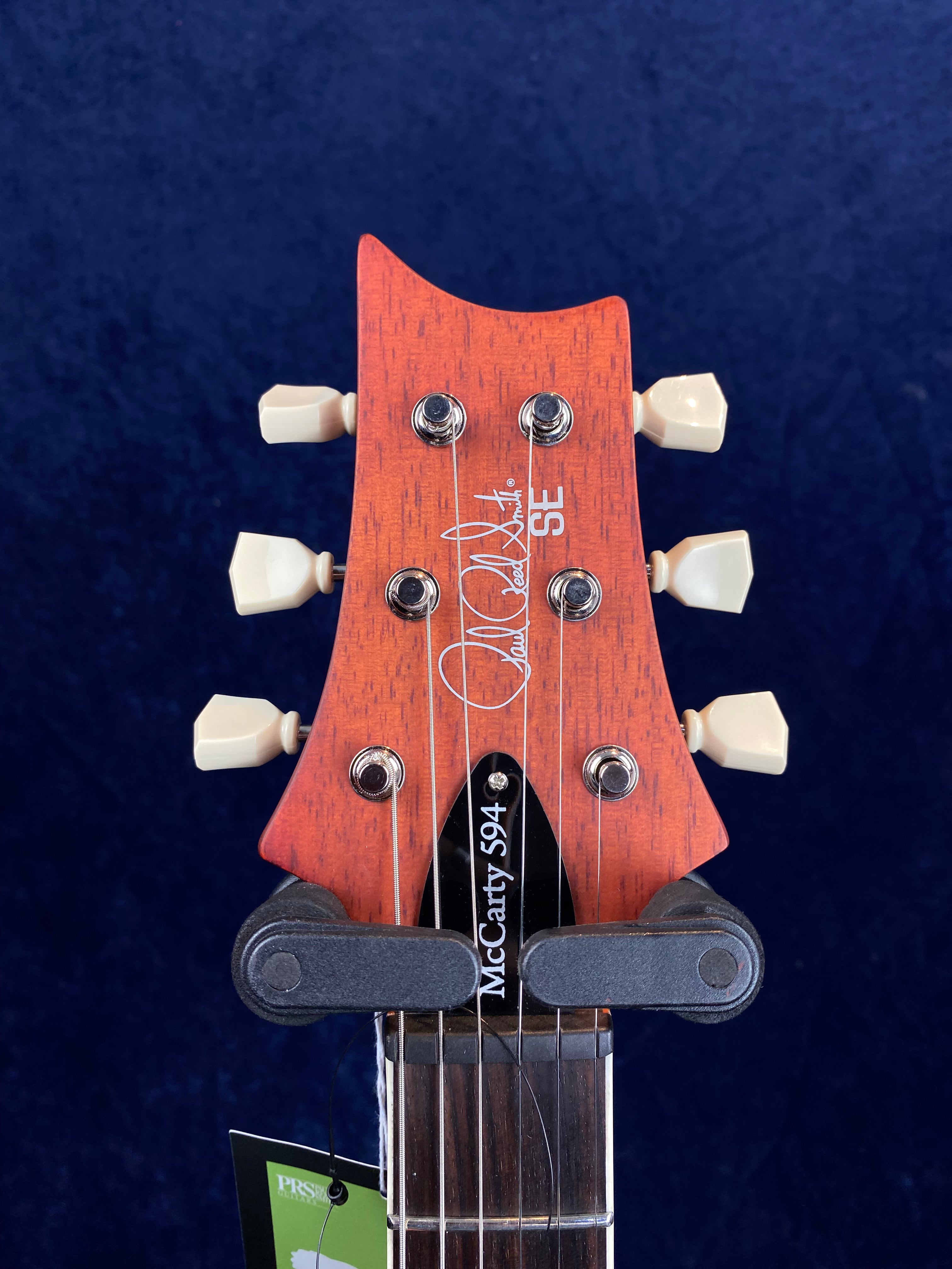 Headstock of a PRS SE electric guitar, orange wood finish, six cream tuners, “McCarty 594” labeled truss rod cover, signature printed on front.
