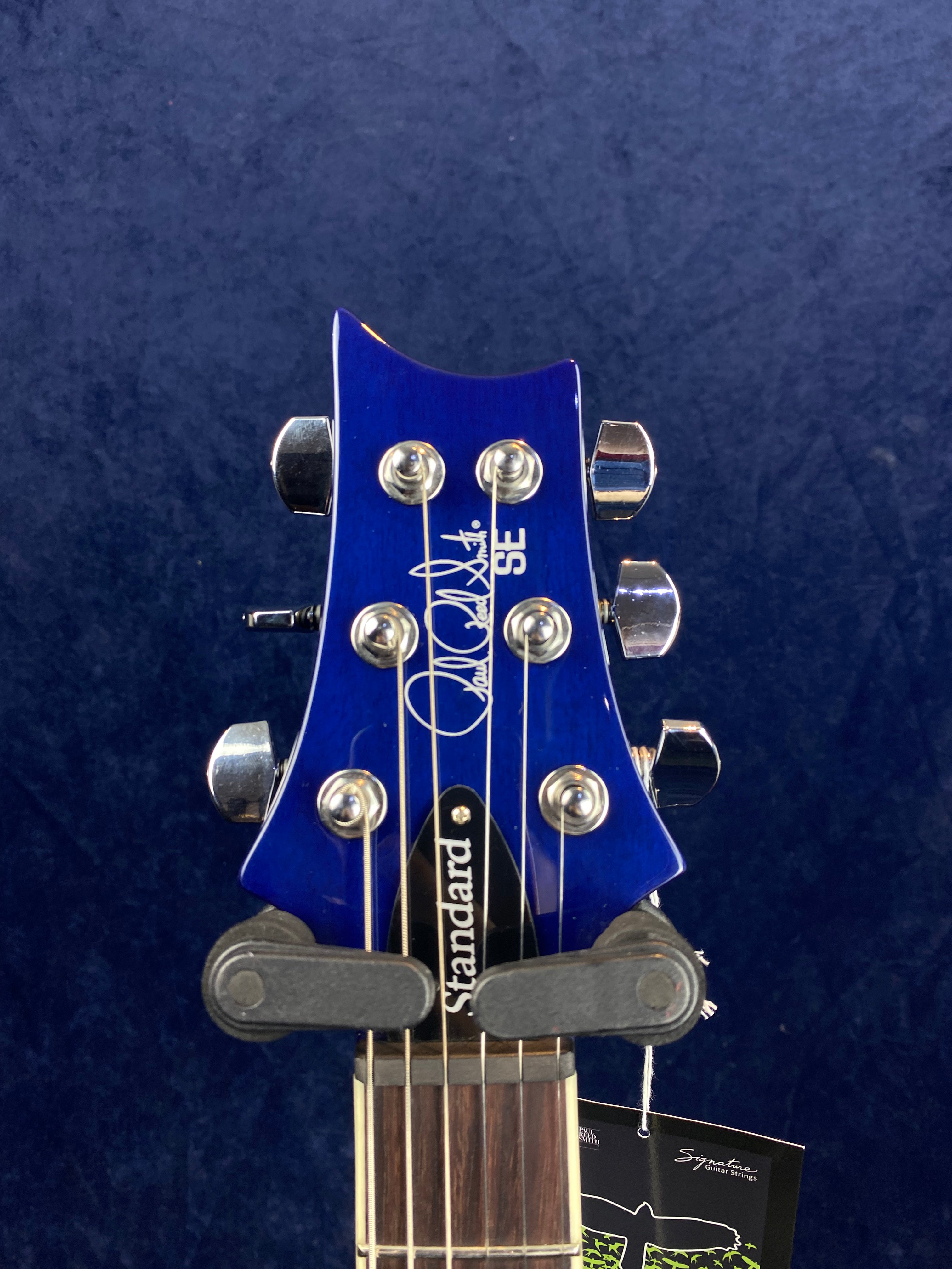 Blue electric guitar headstock with six silver tuning pegs, SE script, and “Standard” truss rod cover, strings running down toward fretboard.