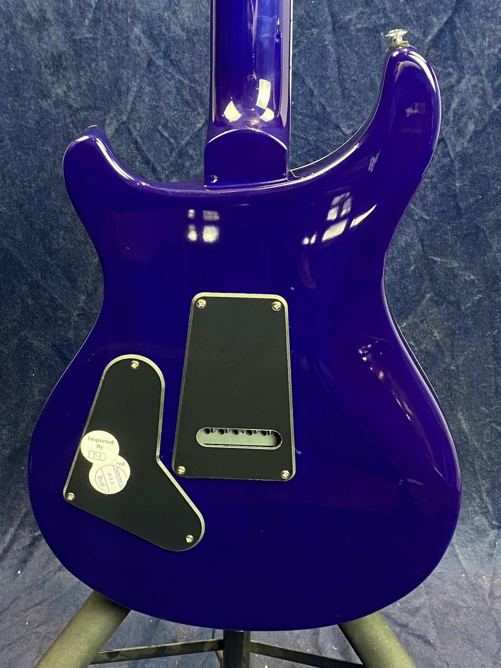 Back view of a glossy purple electric guitar showing two black plastic covers and inspection stickers on the lower plate.