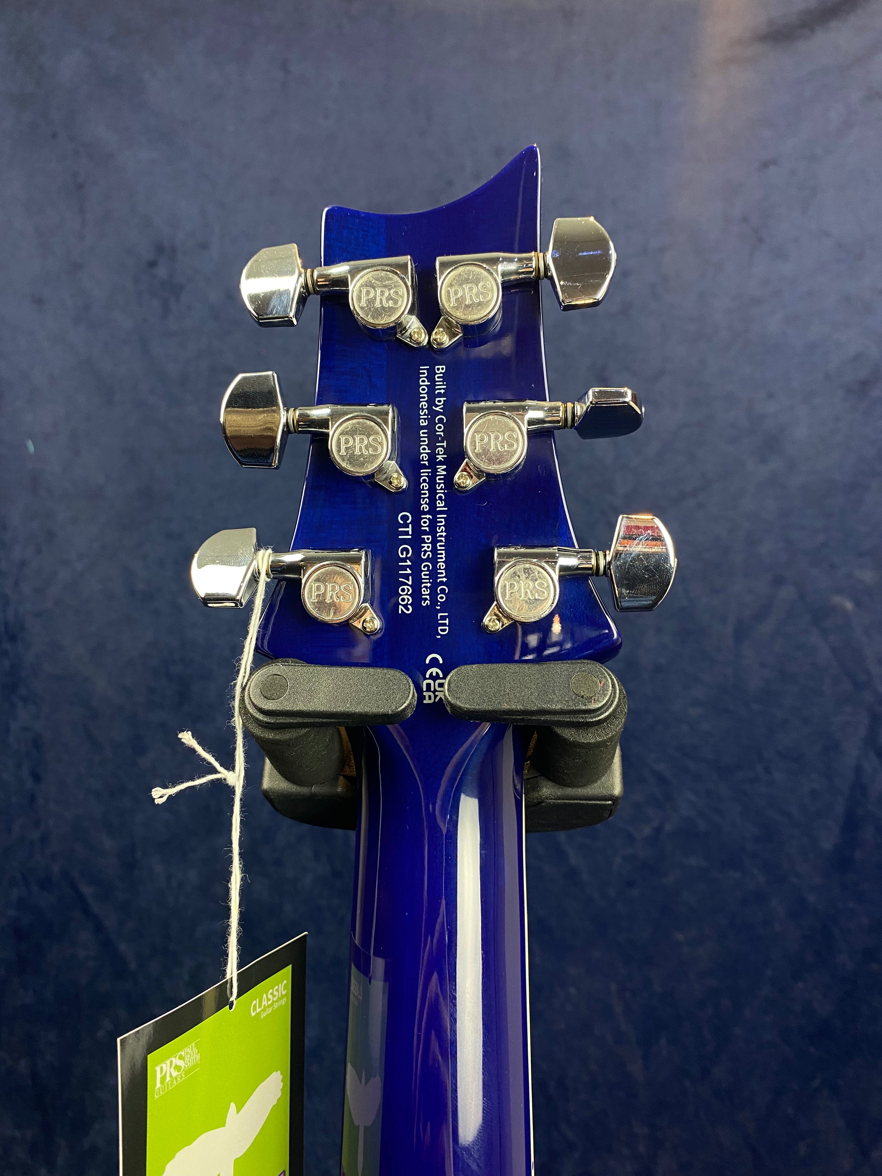 Back of a blue electric guitar headstock with six silver PRS tuning pegs, a white serial number, and a green tag hanging by a string.
