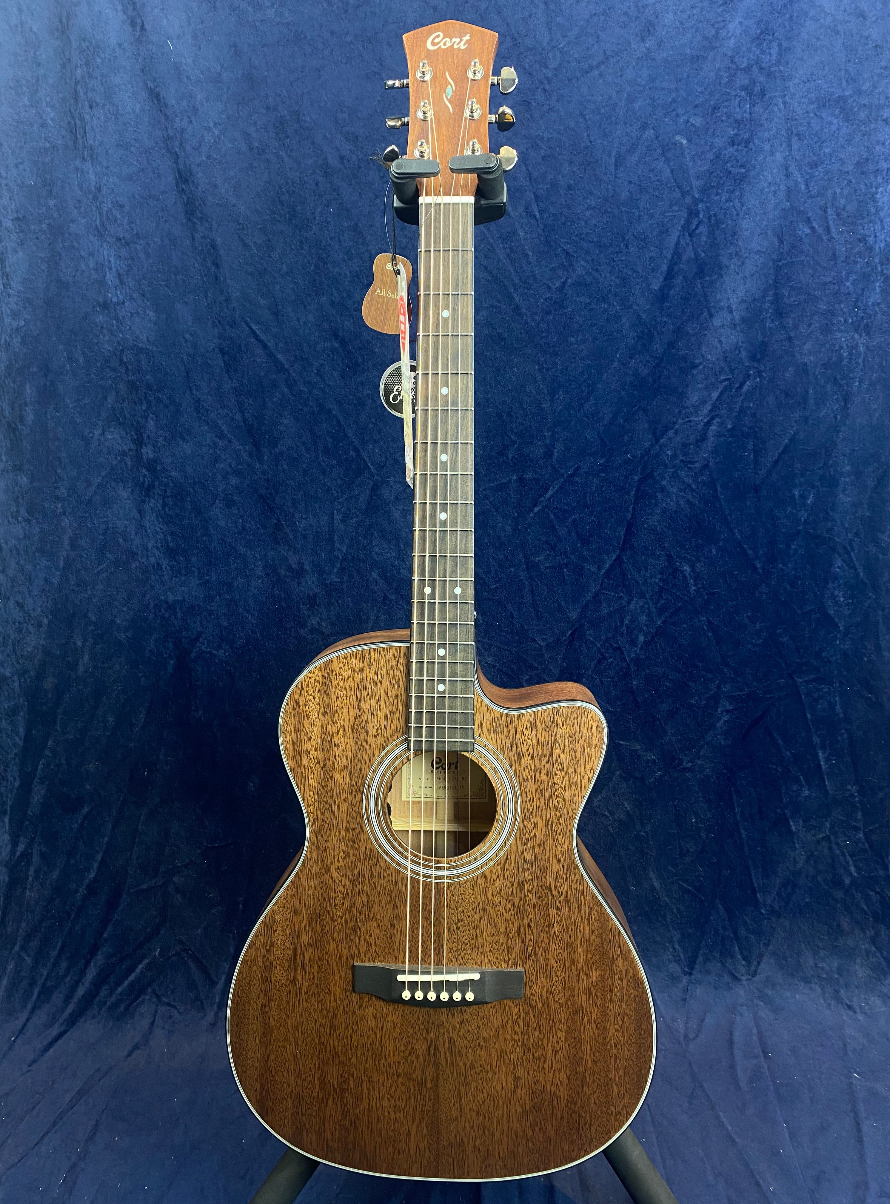 Cort acoustic guitar with a cutaway body, natural wood grain finish, and six strings, displayed upright against a dark blue background.
