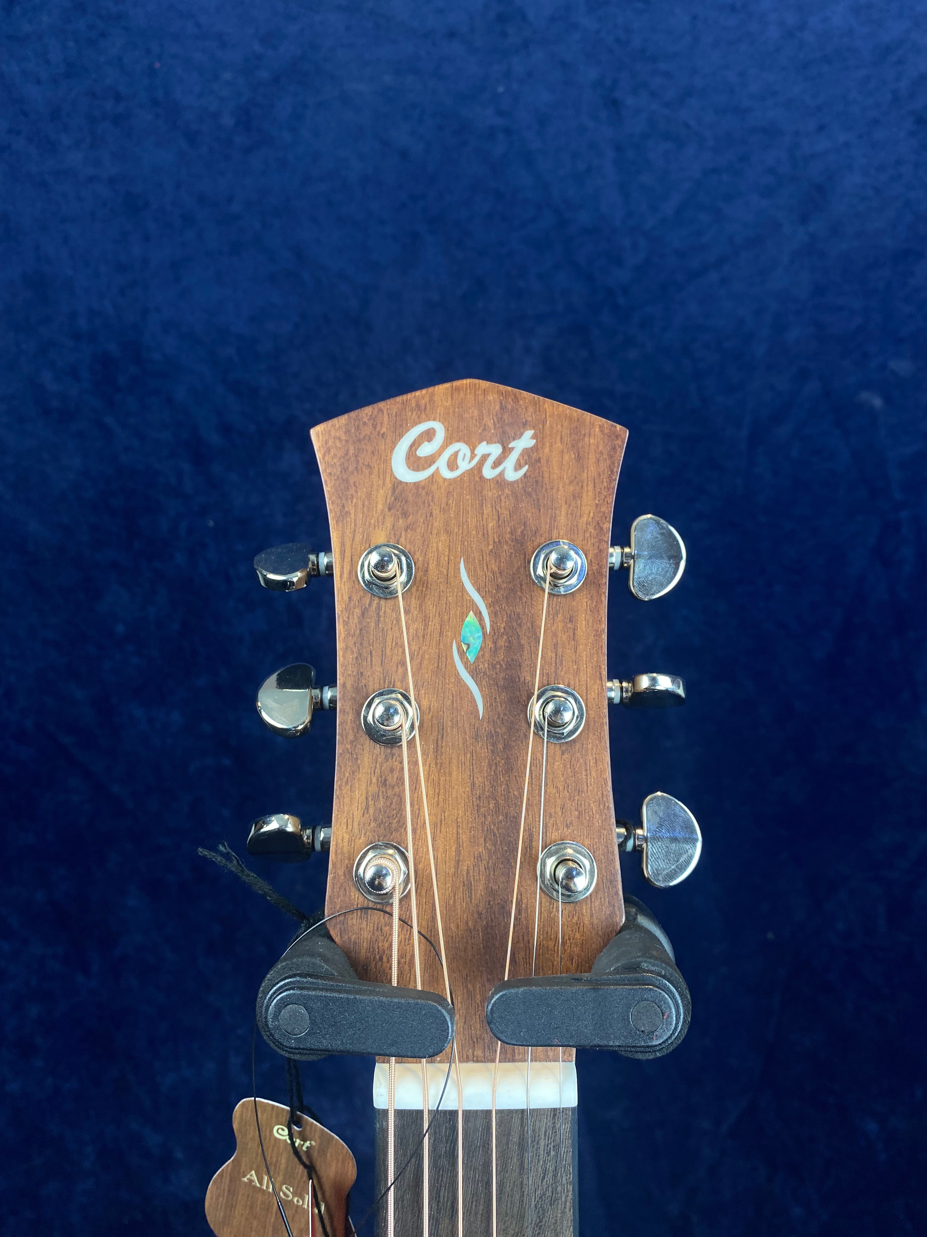 A Cort acoustic guitar headstock with six silver tuning pegs, wood grain finish, and a small decorative inlay below the Cort logo.
