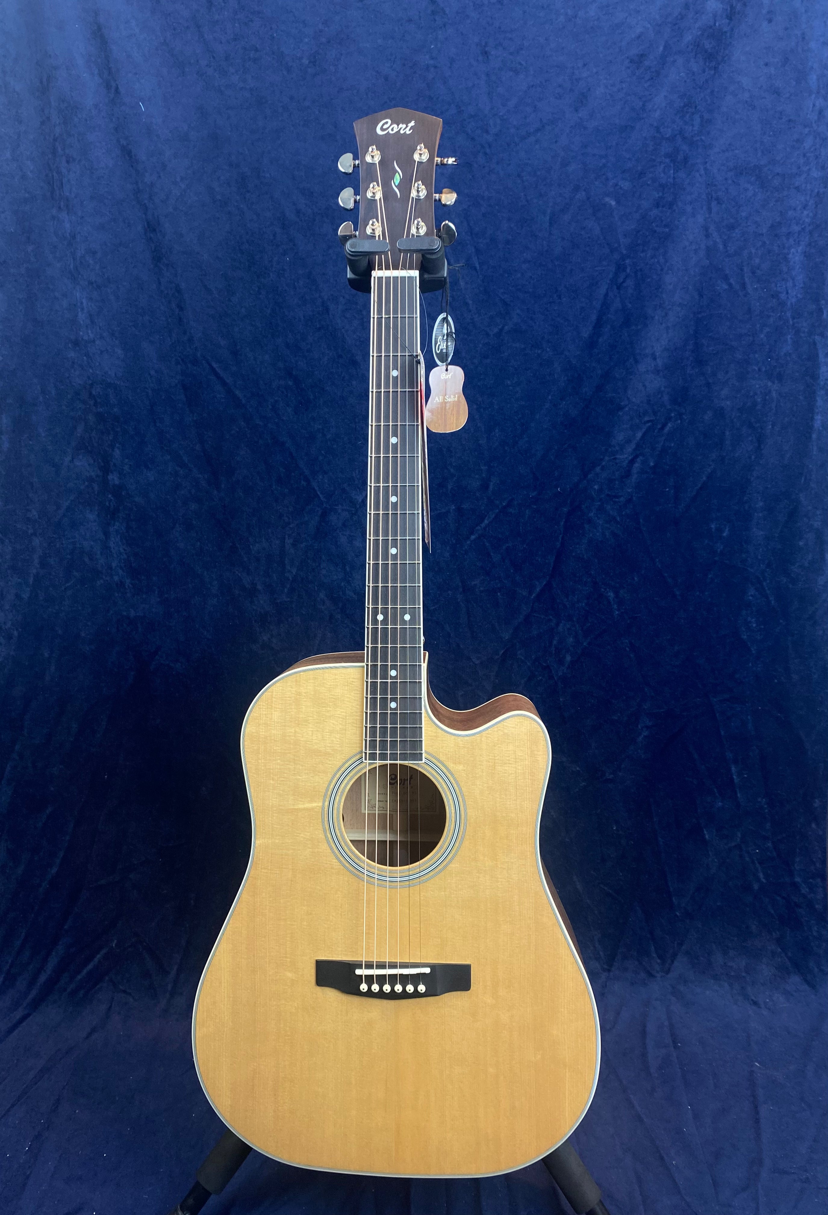 Natural wood acoustic guitar with a single cutaway, dark fretboard, and six strings, standing against a dark blue background.