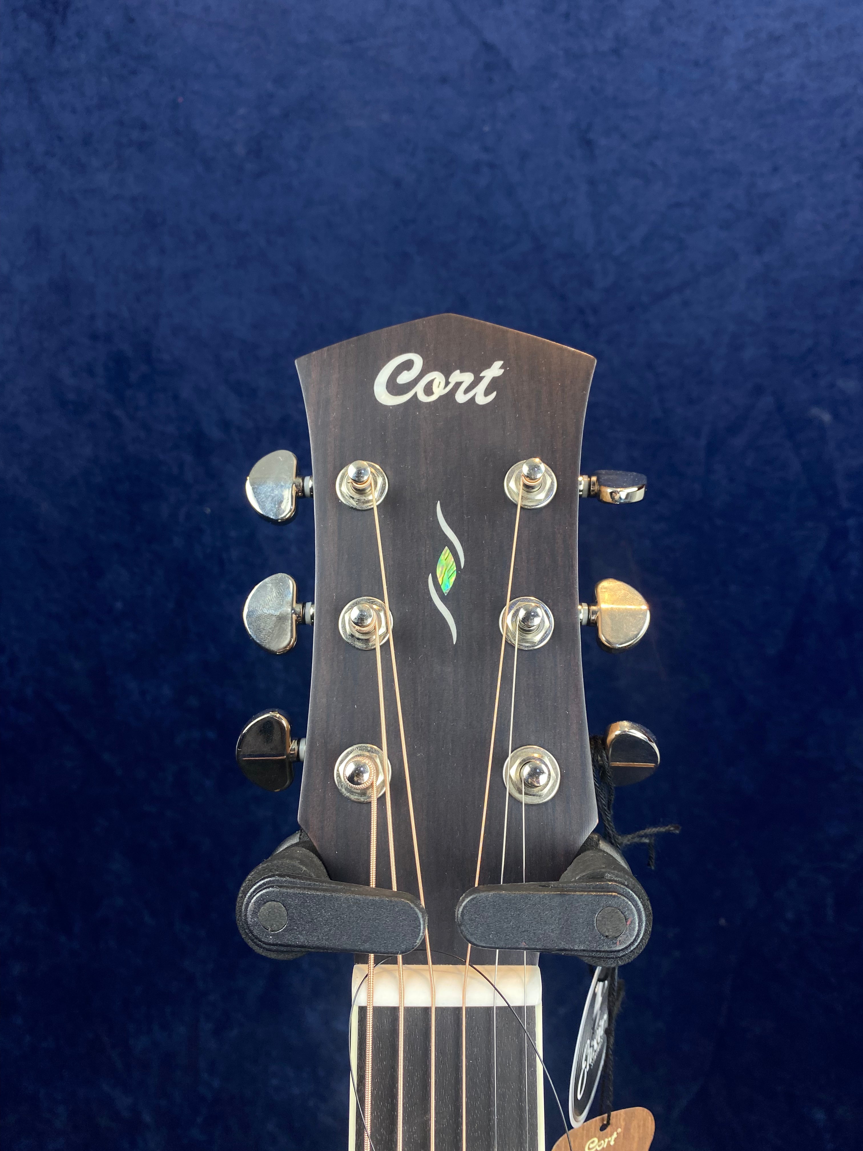 Cort acoustic guitar headstock with six tuning pegs, strings, and a small green inlay below the Cort logo against a dark blue background.
