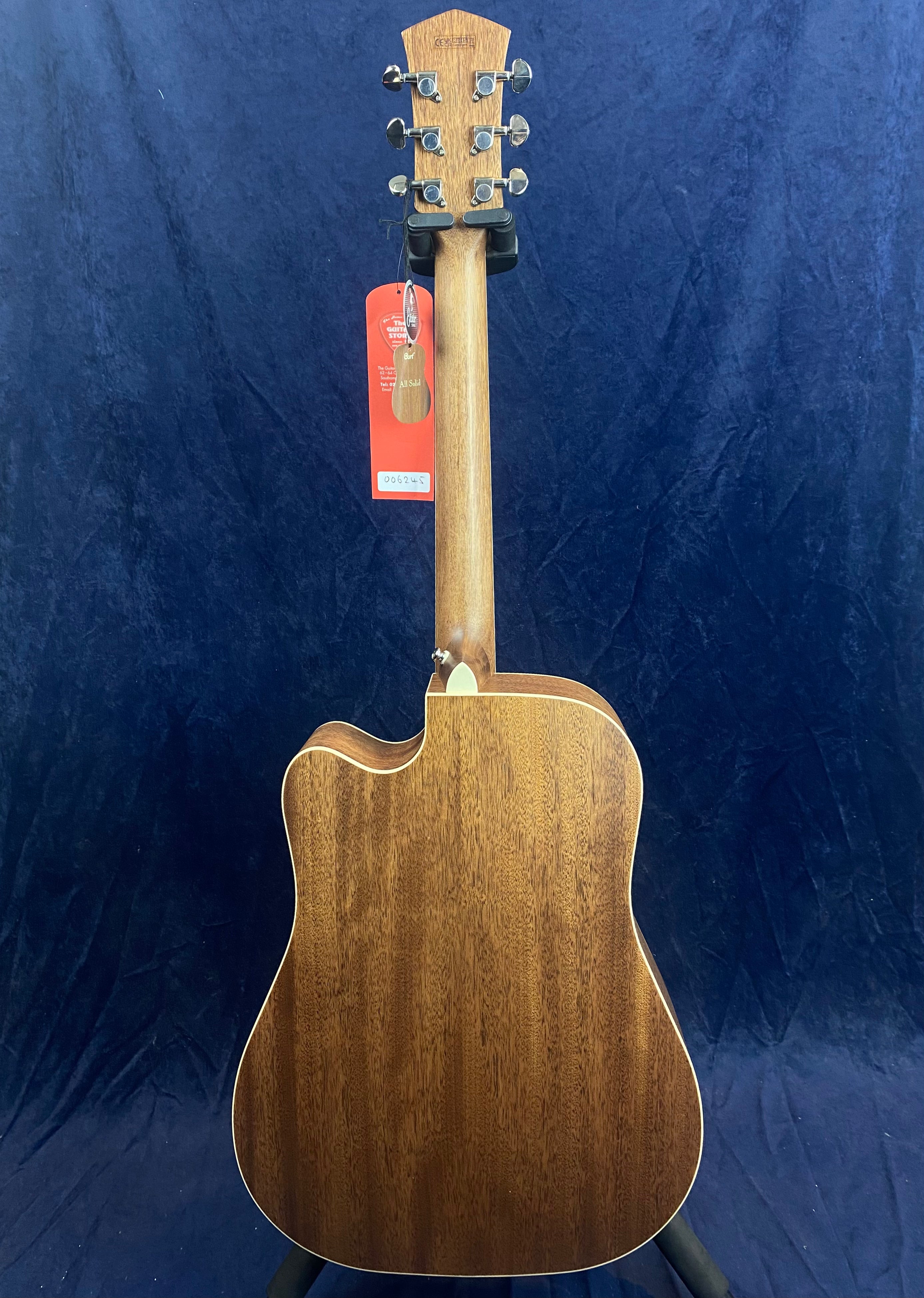 Brown acoustic guitar viewed from the back, showing wood grain, light trim, a neck heel, and a red tag hanging from the headstock.
