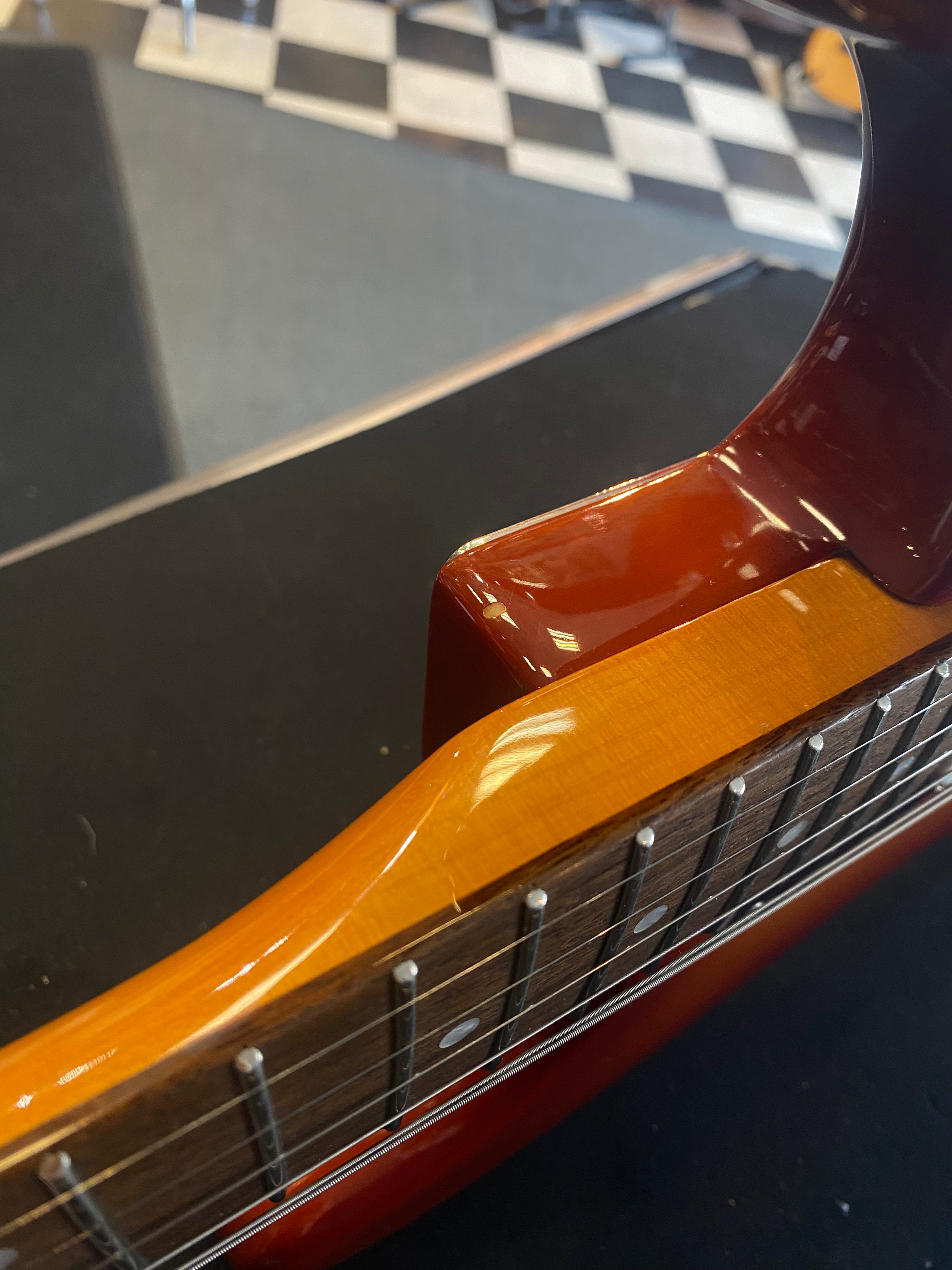 Close-up of an electric guitar’s neck joint, showing glossy wood finish, dot inlays, and six strings; a black and white tile floor is in the background.