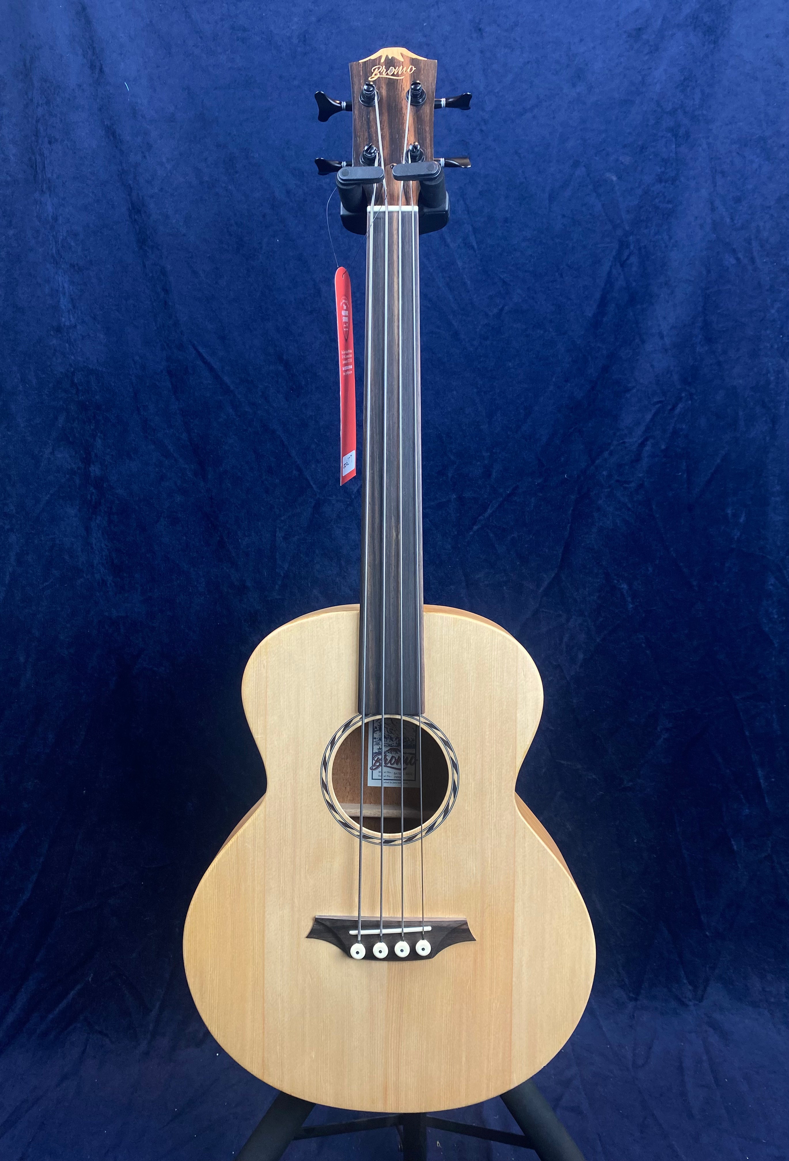 Acoustic fretless bass guitar with a natural wood finish, four strings, round sound hole, and a dark wood headstock against a dark blue background.