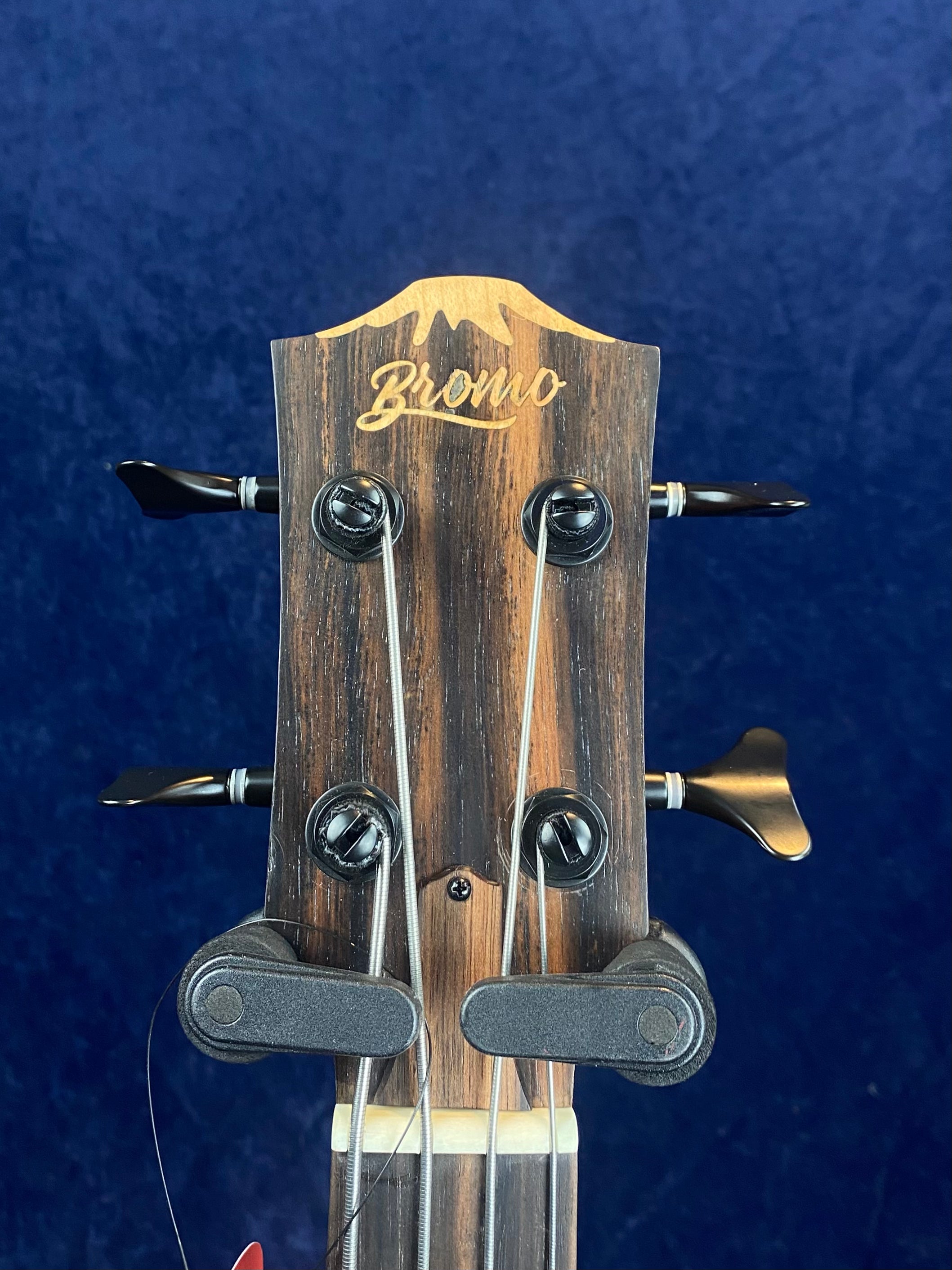 Close-up of a Bromo bass guitar headstock, four tuning pegs, two visible string retainers, wood grain finish, and "Bromo" logo at the top.