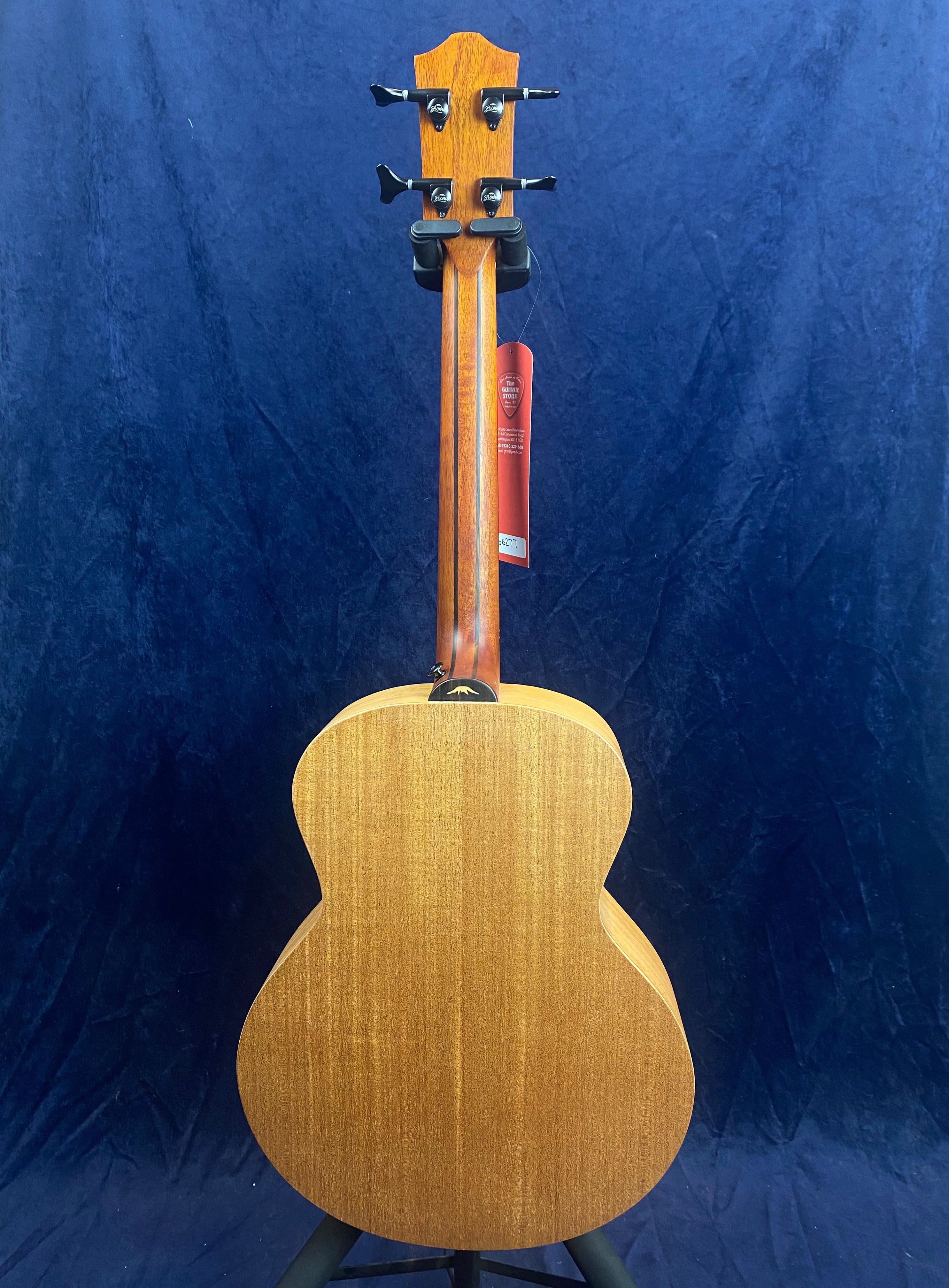 The back of a wooden bass guitar with a natural finish, four tuning pegs, and a red tag hanging from the headstock, seen against a dark blue background.