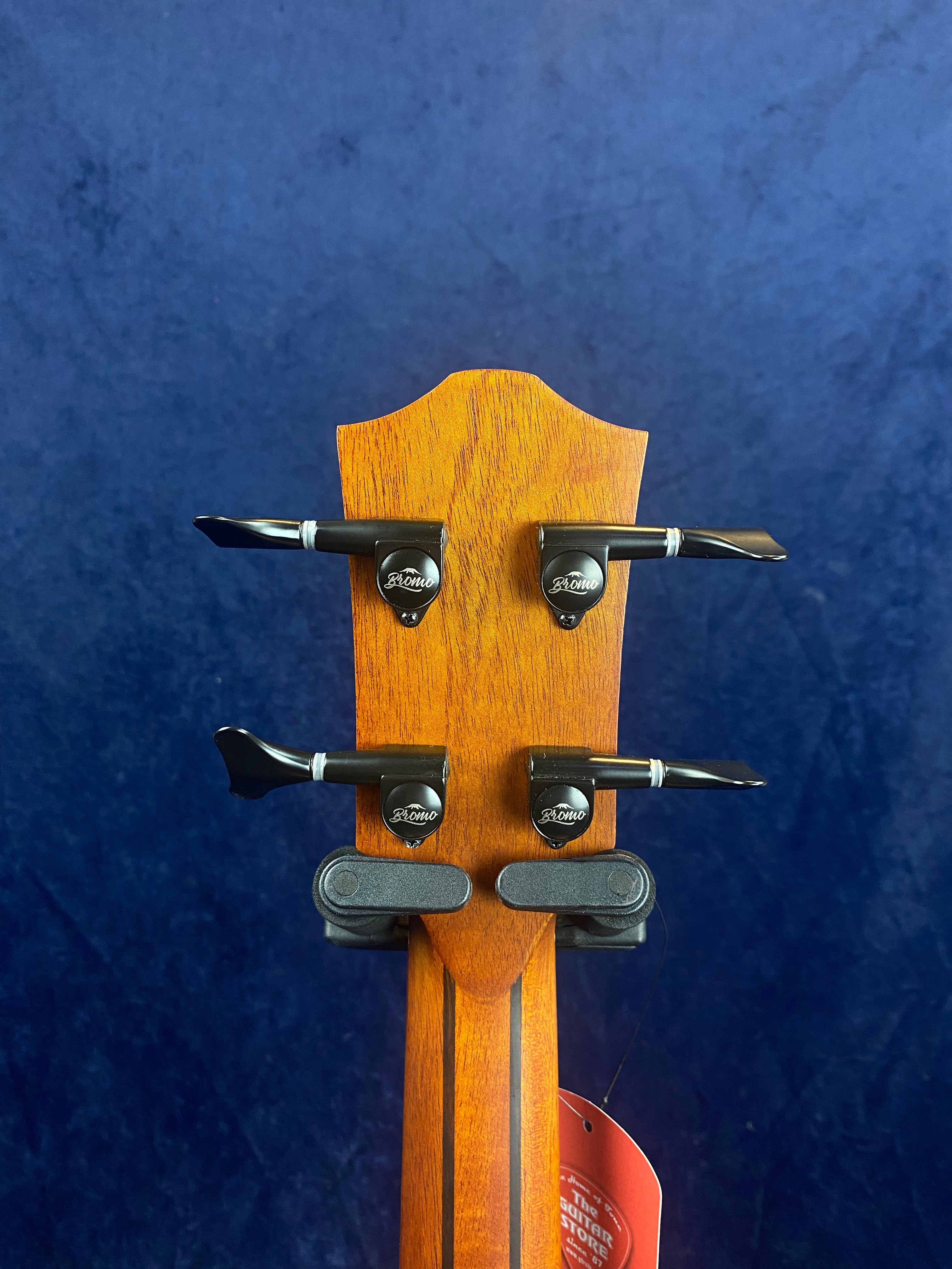 Back of a bass guitar headstock with four black tuning pegs, displaying the brand name “Bromo,” against a dark blue background.