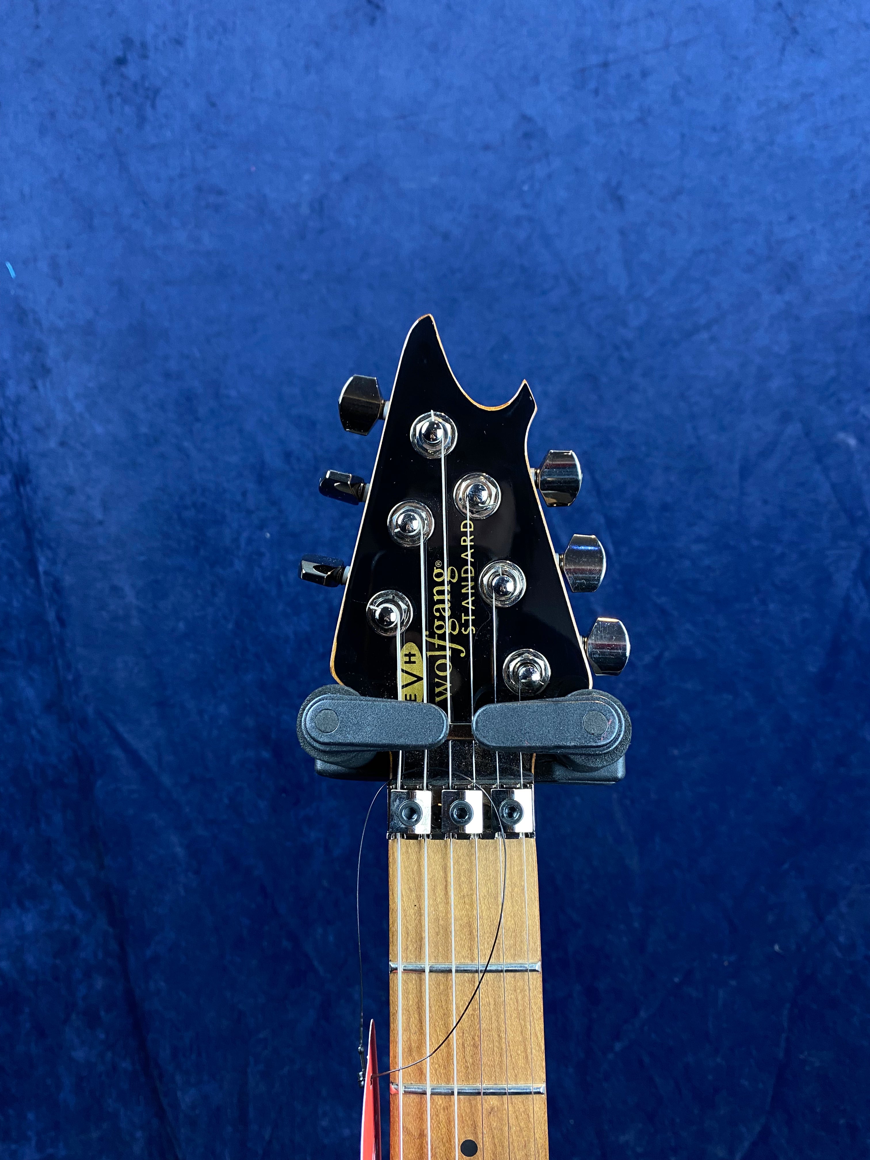 Electric guitar headstock with six tuning pegs, maple fretboard, string retainer bars, and "Wolfgang" branding against a blue background.