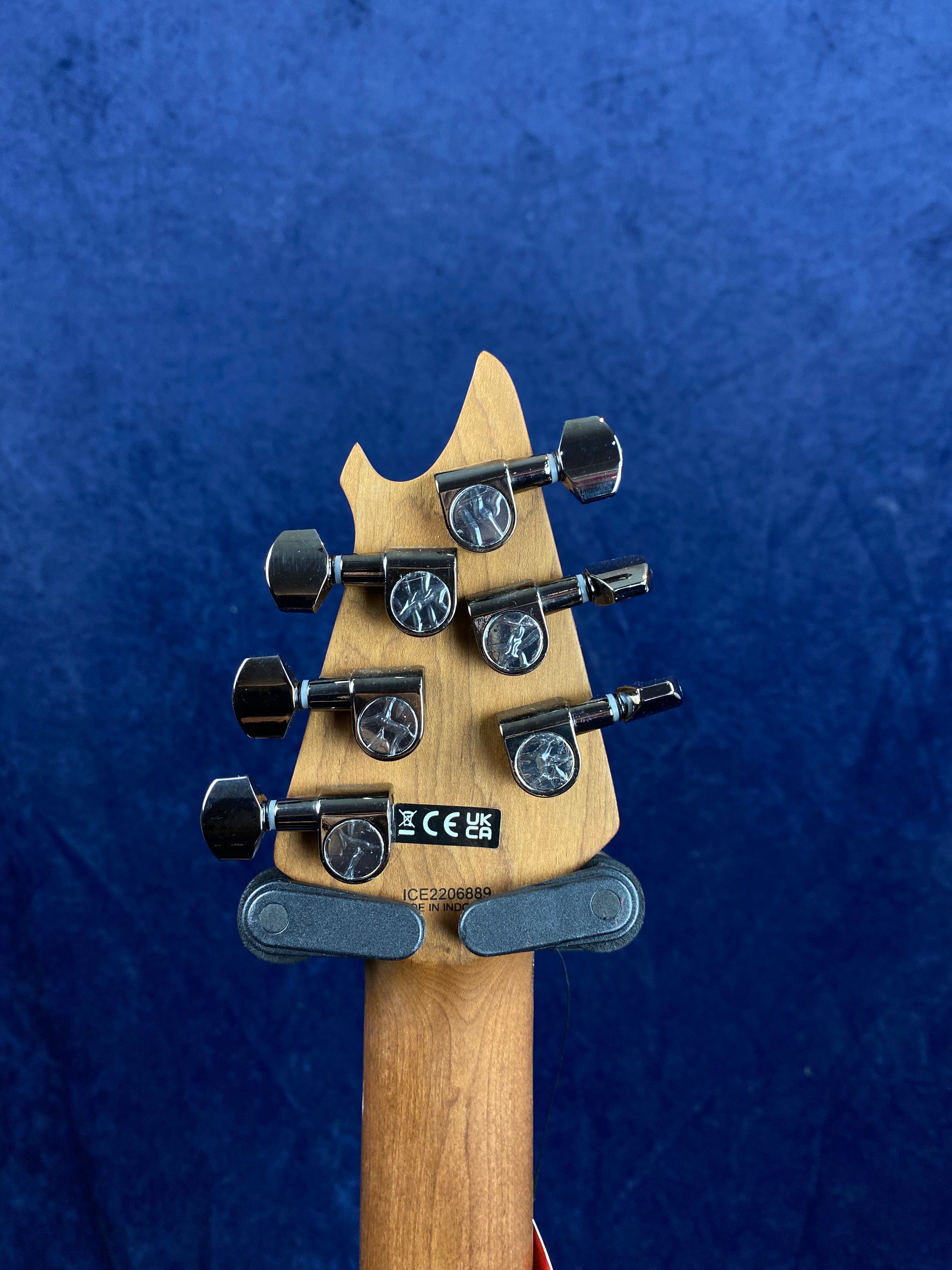 Six chrome tuning pegs on a natural wood electric guitar headstock, viewed from the back, with a blue background and certification stickers visible.