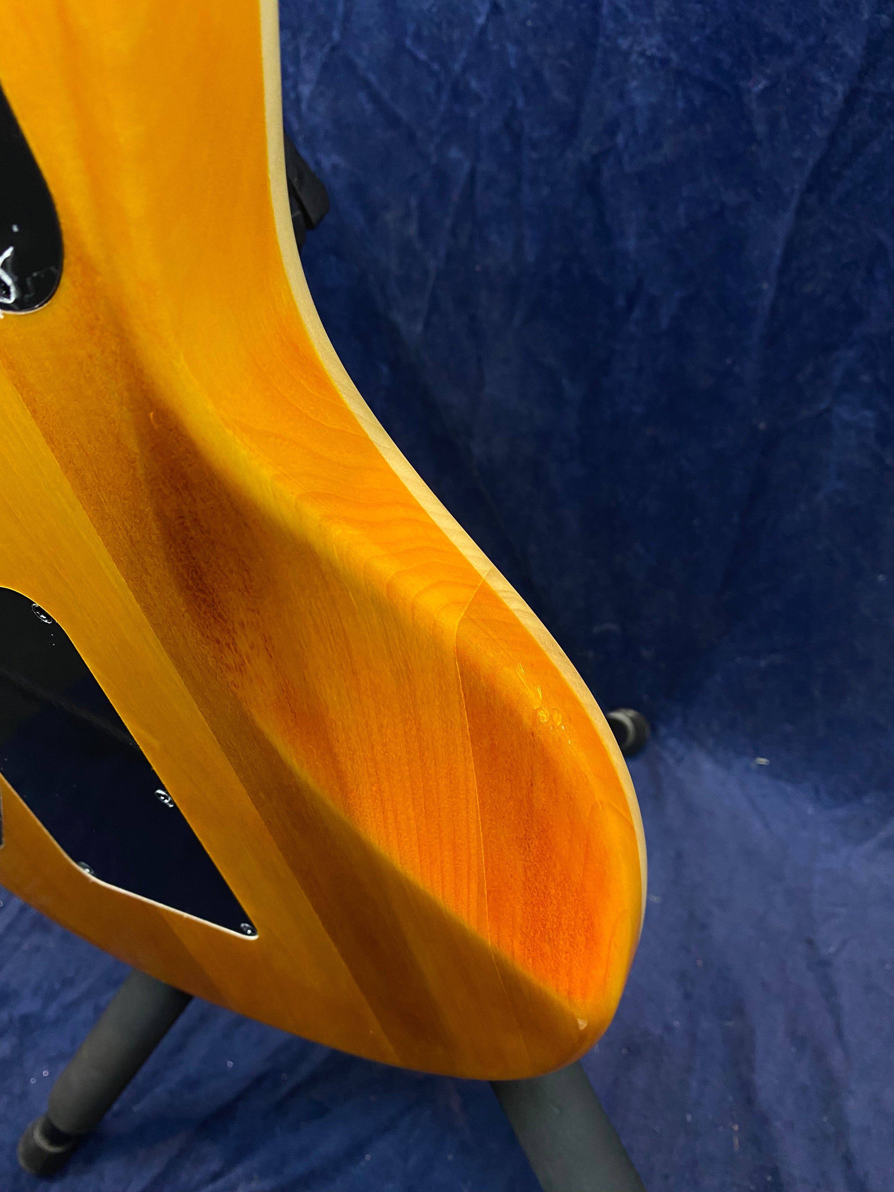 Glossy orange and yellow sunburst electric guitar body on a stand, showing smooth contours and a black plastic back plate against a blue background.