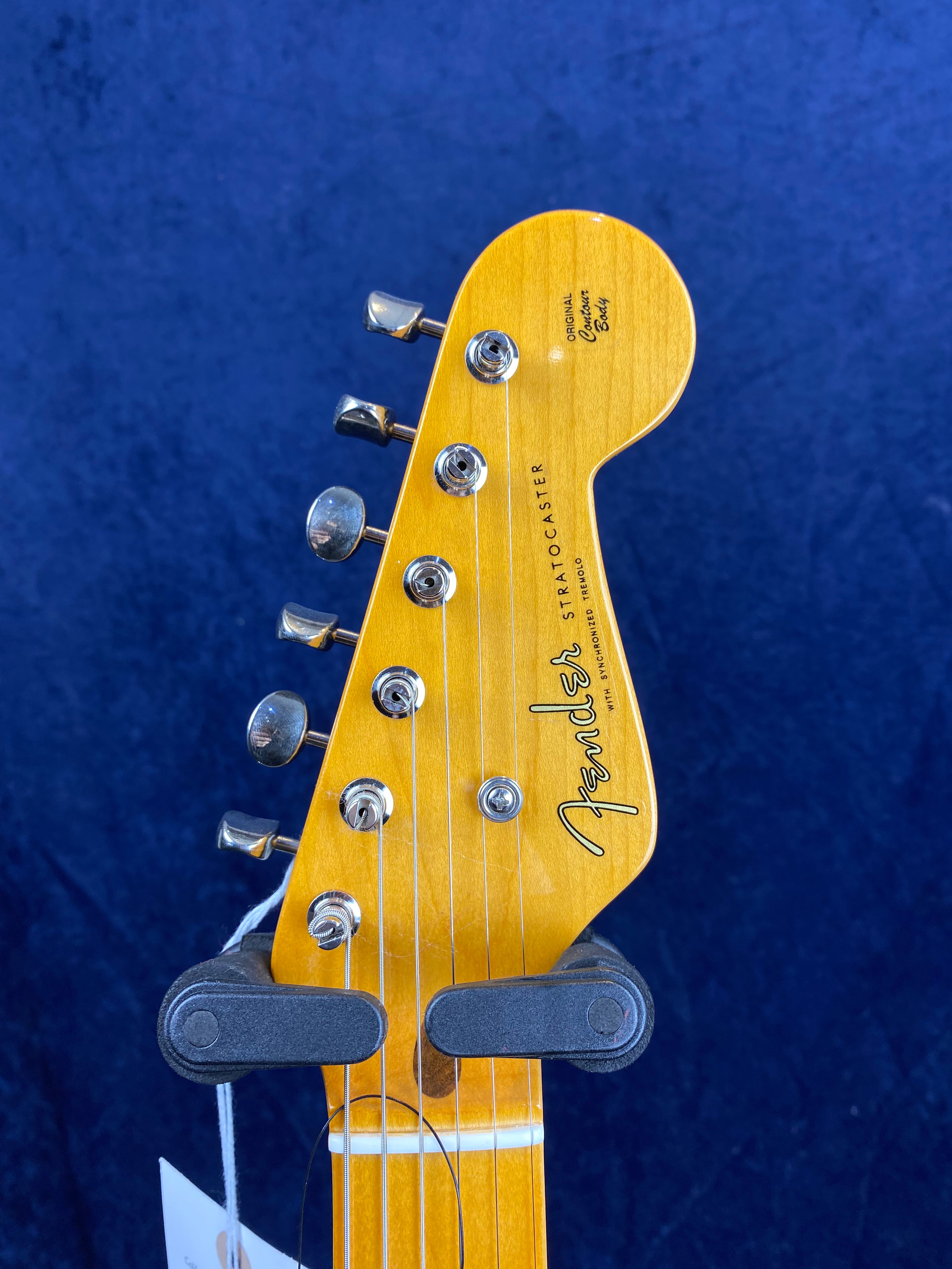 Light wood Fender Stratocaster electric guitar headstock with six chrome tuning pegs, black logo, and visible strings against a dark blue background.