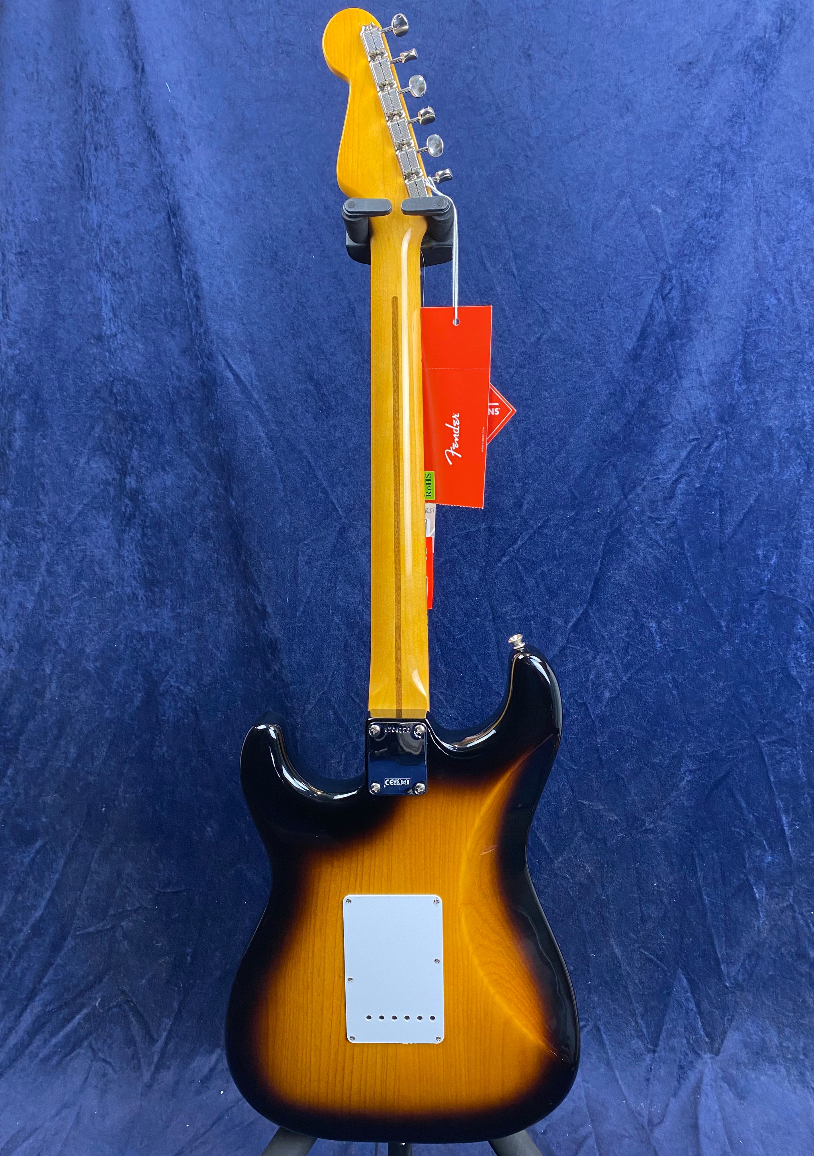 Sunburst Fender electric guitar shown from the back, maple neck, chrome tuners, white backplate, and red Fender tag hanging from a tuning peg.