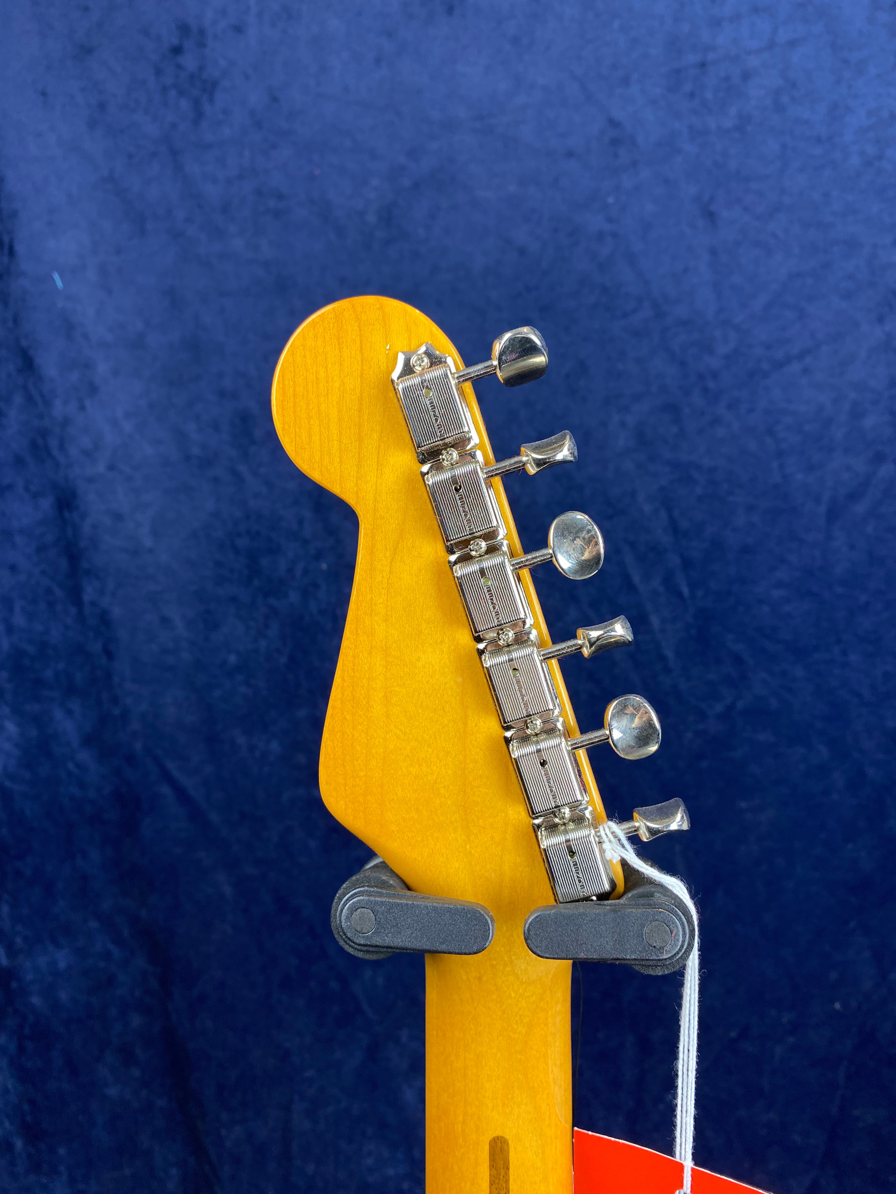 Six silver tuning pegs on a light wood electric guitar headstock, held by a wall mount against a deep blue background.