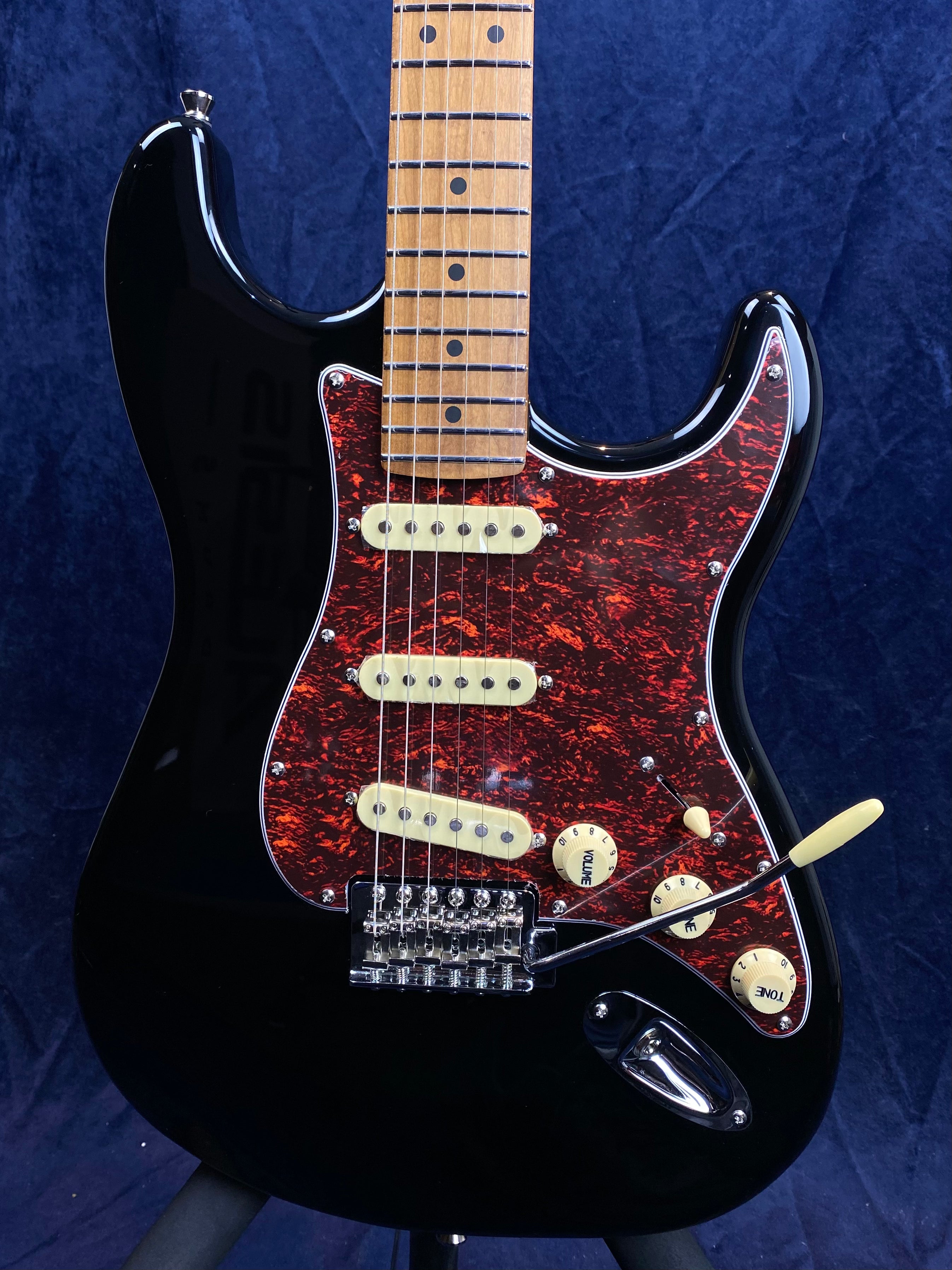 Black electric guitar with a maple neck, red tortoiseshell pickguard, three cream single-coil pickups, and cream control knobs.