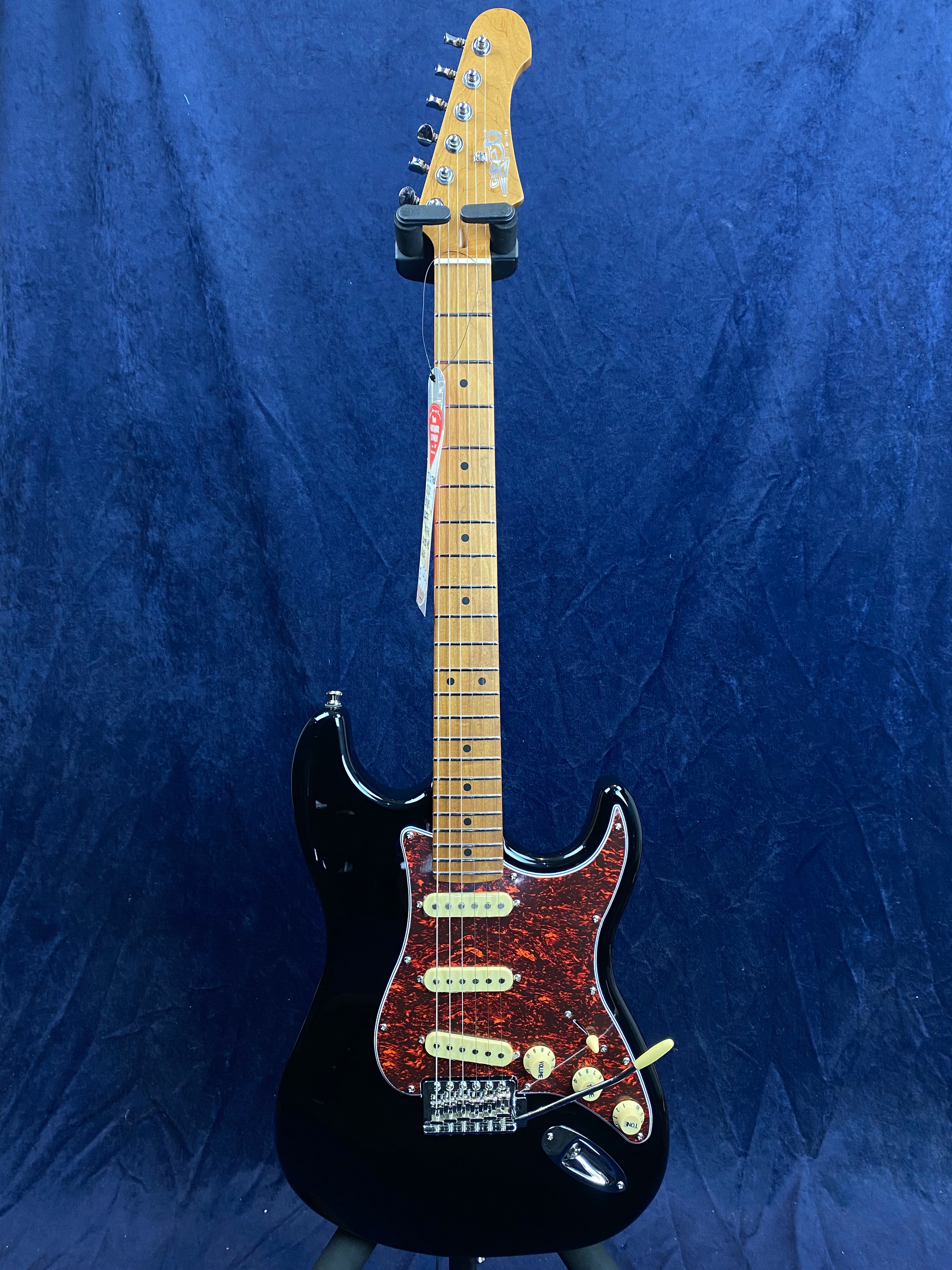 Black electric guitar with a maple neck, three cream single-coil pickups, and a red tortoiseshell pickguard, displayed upright against a blue backdrop.