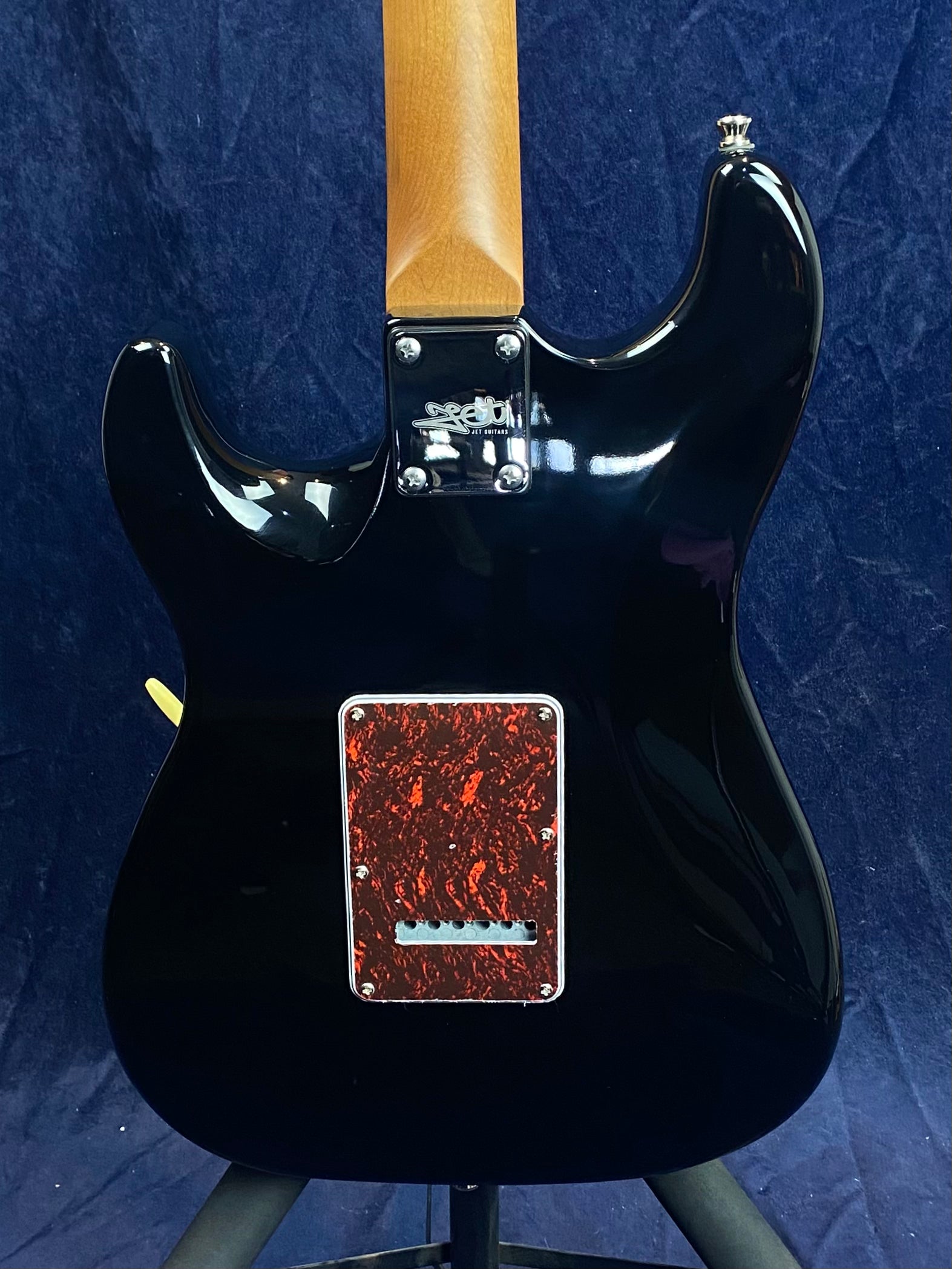 Black electric guitar viewed from the back, featuring a brown neck and a red marbled rectangular cover plate near the base.