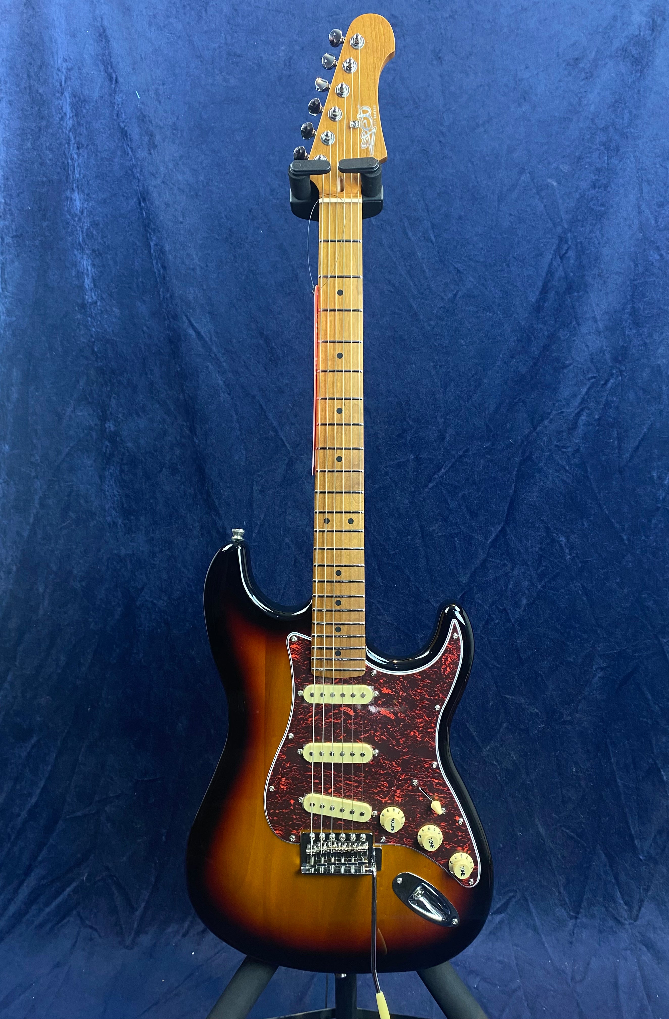 Sunburst electric guitar with a maple neck, three single-coil pickups, cream knobs, and a red tortoiseshell pickguard, shown upright on a stand.