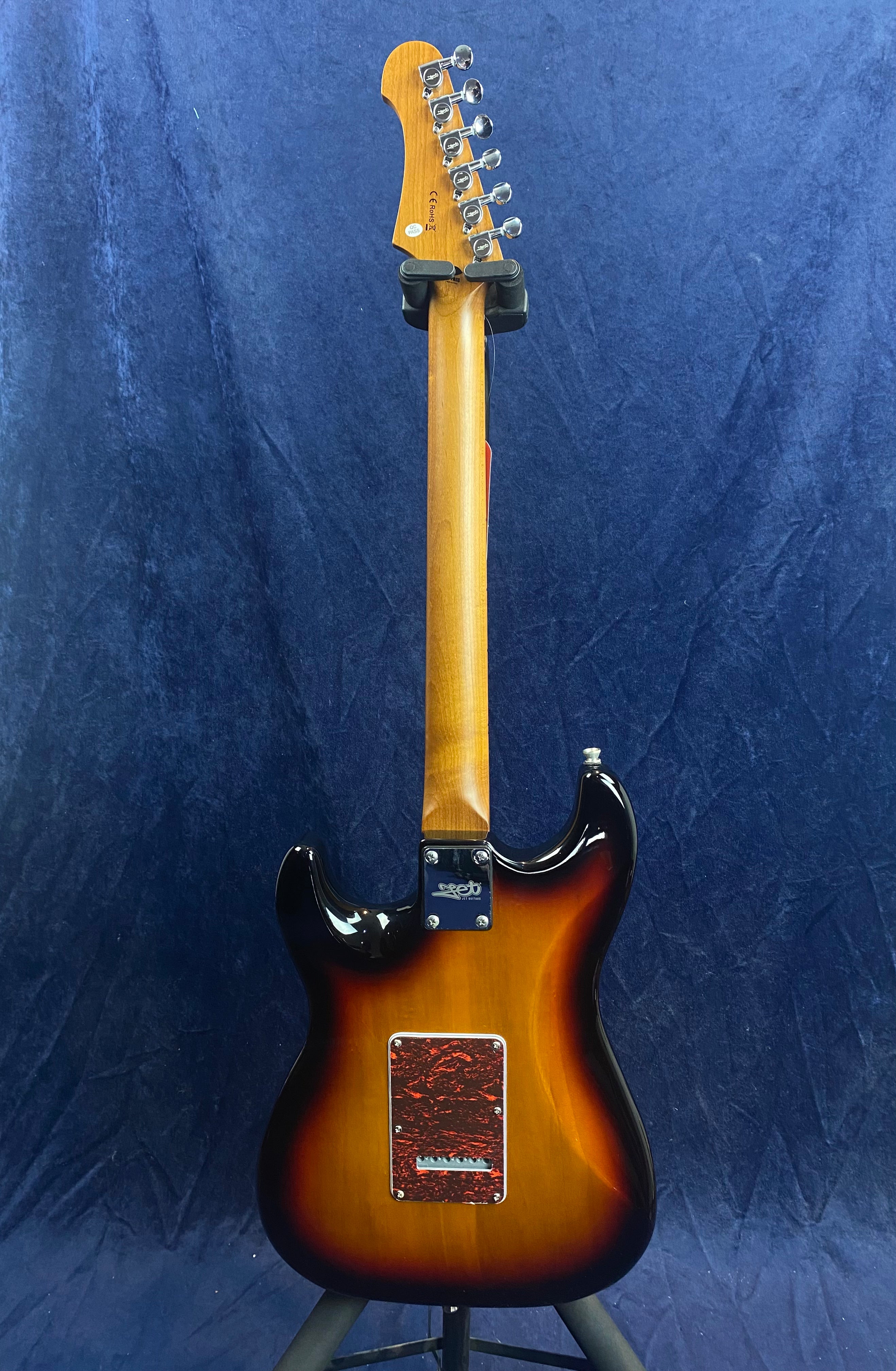 Sunburst electric guitar shown from the back, with a natural wood neck, chrome tuners, and a red marbled backplate against a dark blue background.