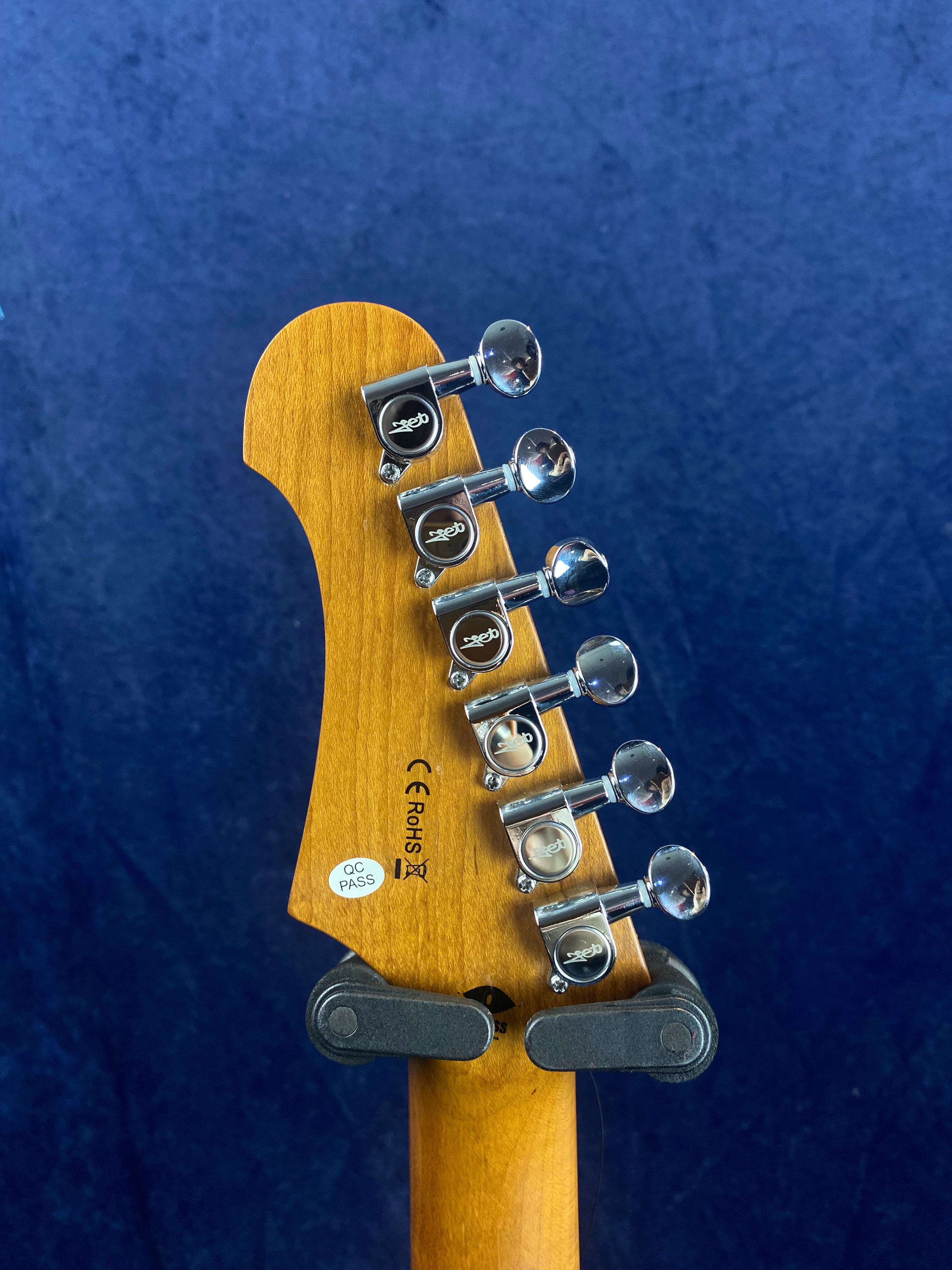 Six chrome tuning pegs line the back of a light wood electric guitar headstock, marked with inspection and certification stickers, against a blue background.