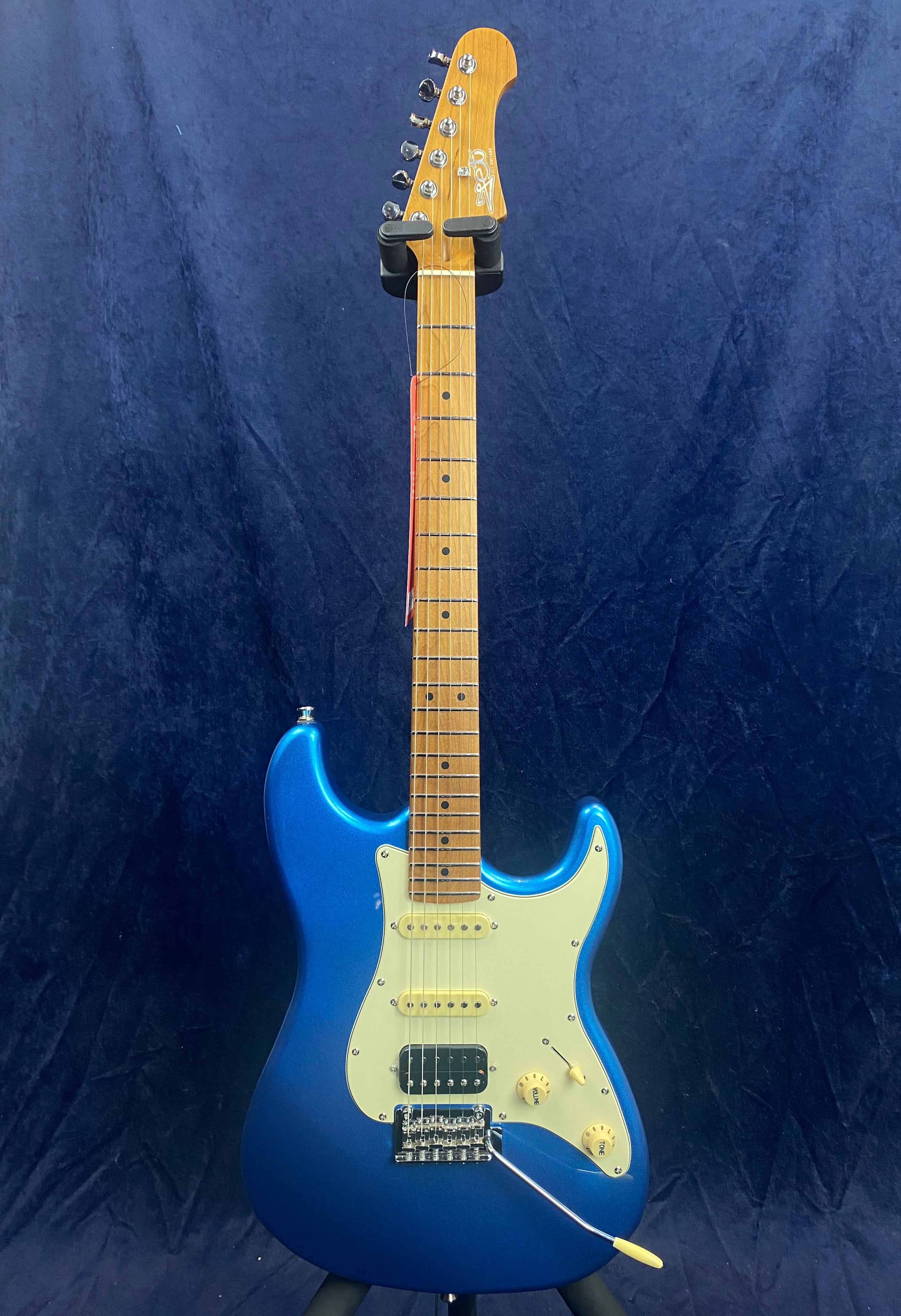Metallic blue electric guitar with a white pickguard, maple neck, three pickups, white knobs, and a tremolo bar, standing upright against a dark background.