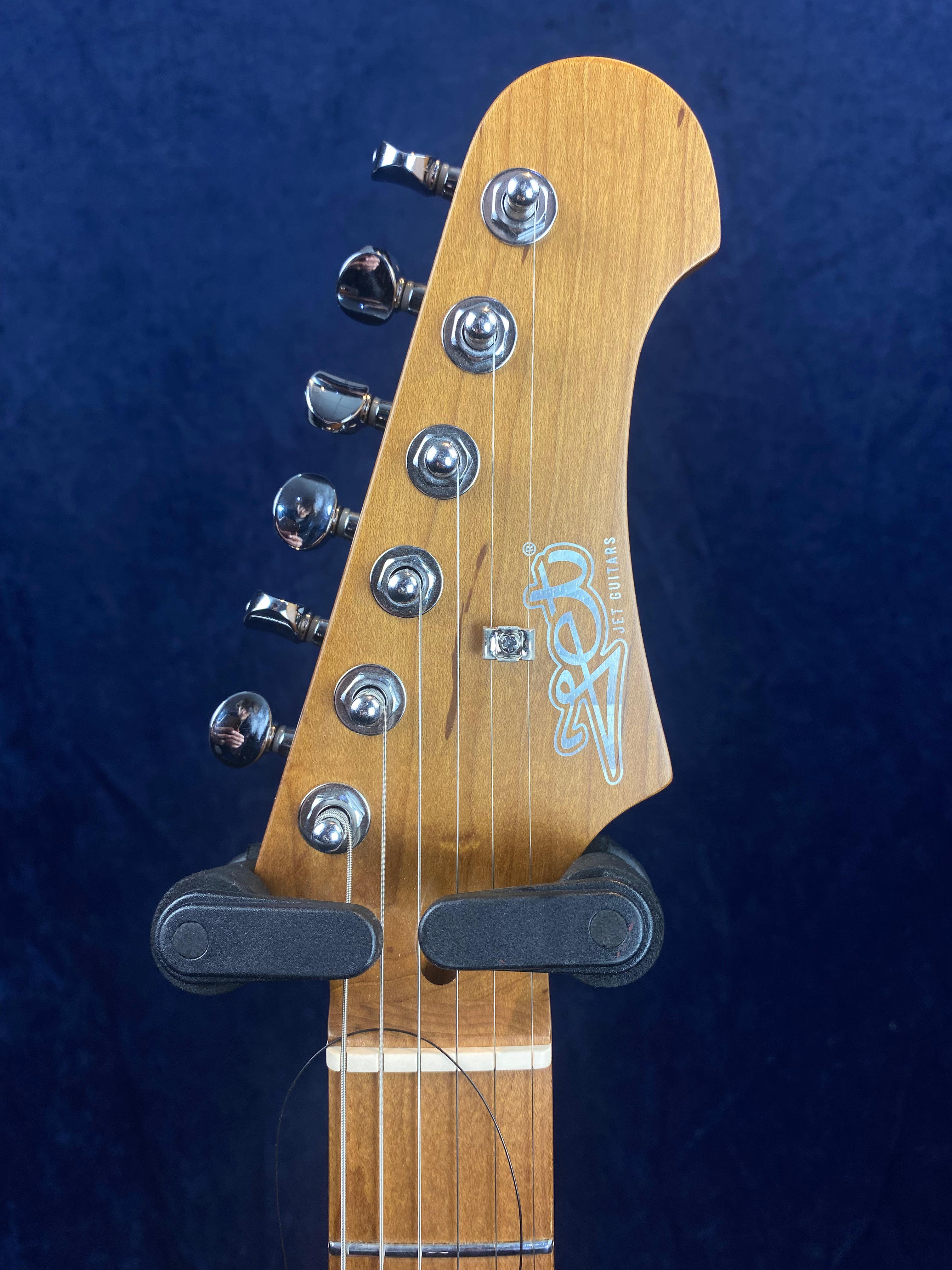 Six metal tuning pegs on a natural wood electric guitar headstock, with the Jet Guitars logo in white and a black capo attached below the pegs.