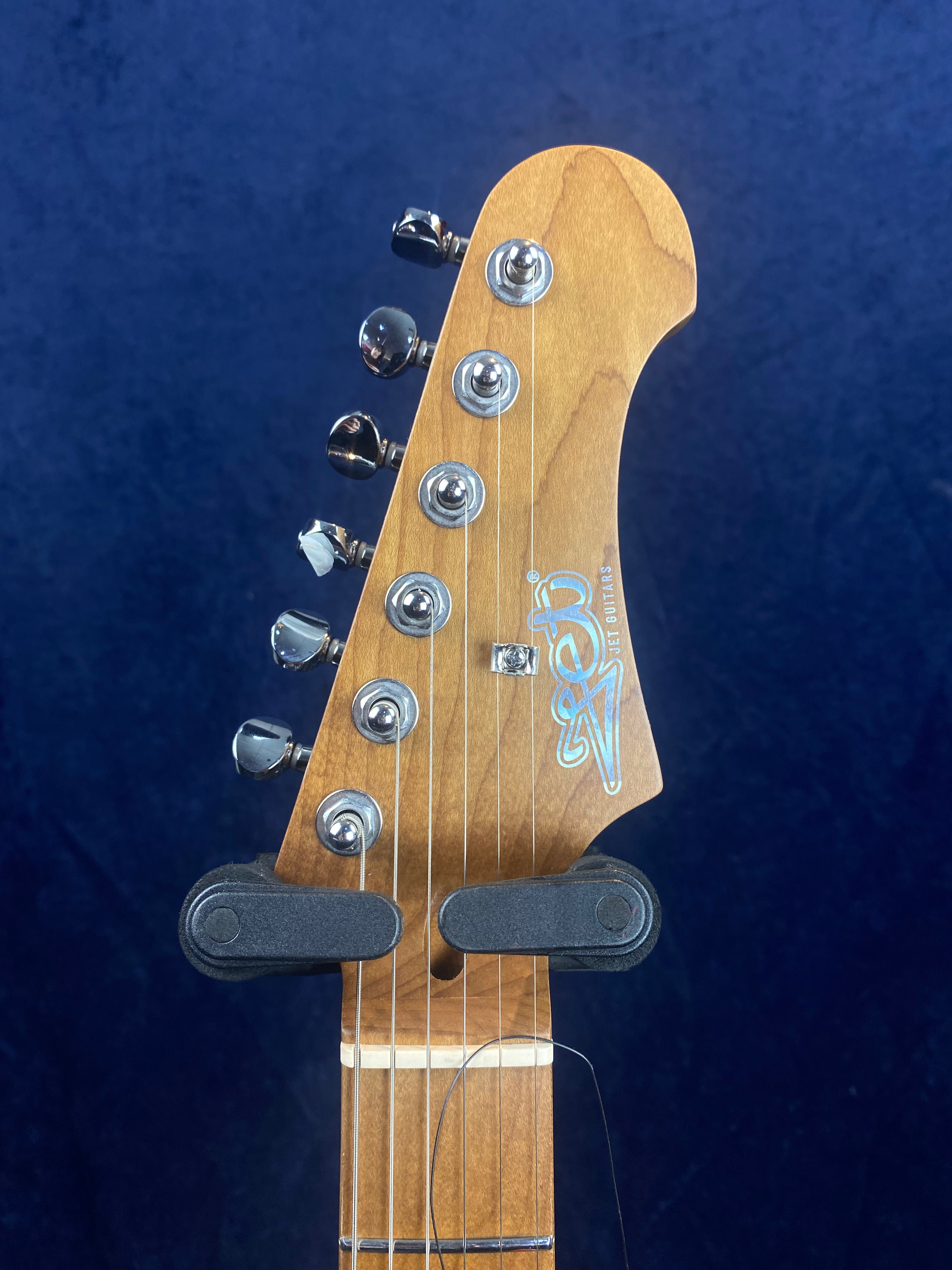 Six-string electric guitar headstock with chrome tuning pegs, a wooden finish, and a logo reading "Zeq" in light blue.