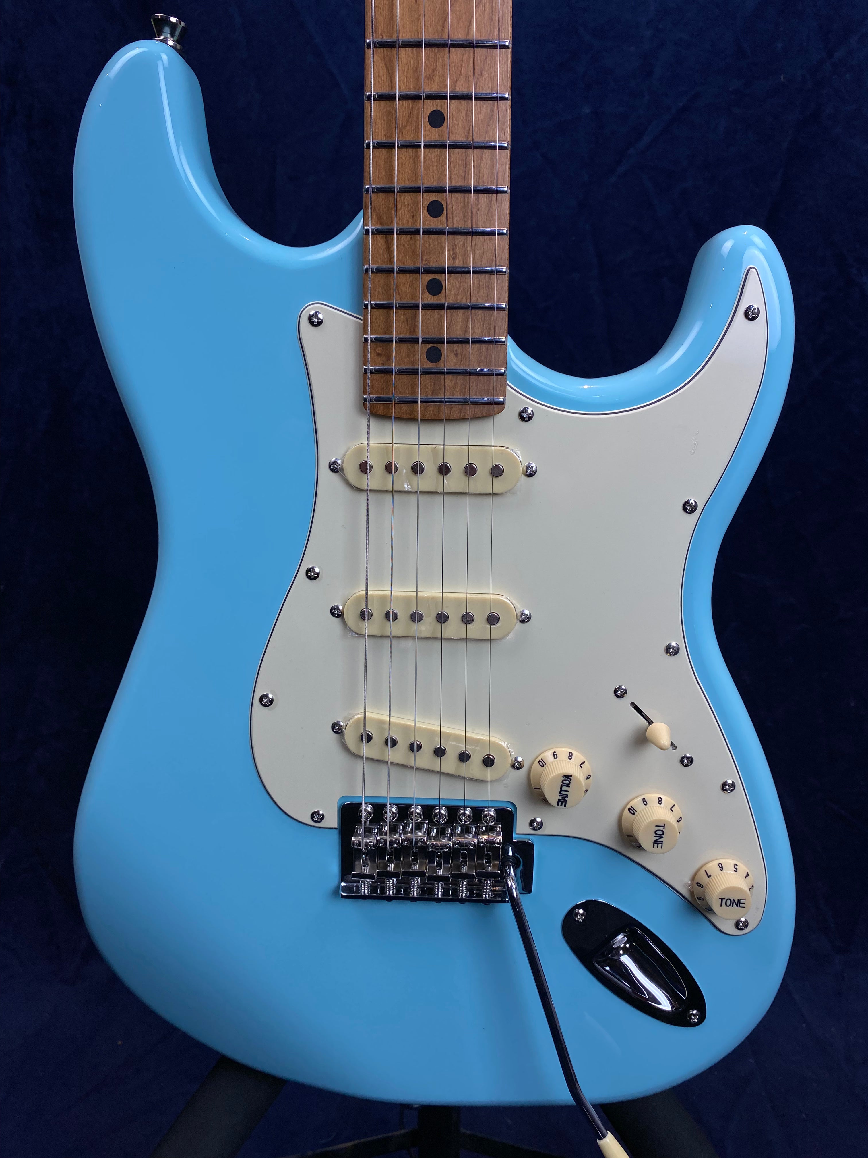 Light blue electric guitar with a white pickguard, three single-coil pickups, and three cream-colored control knobs on a dark background.