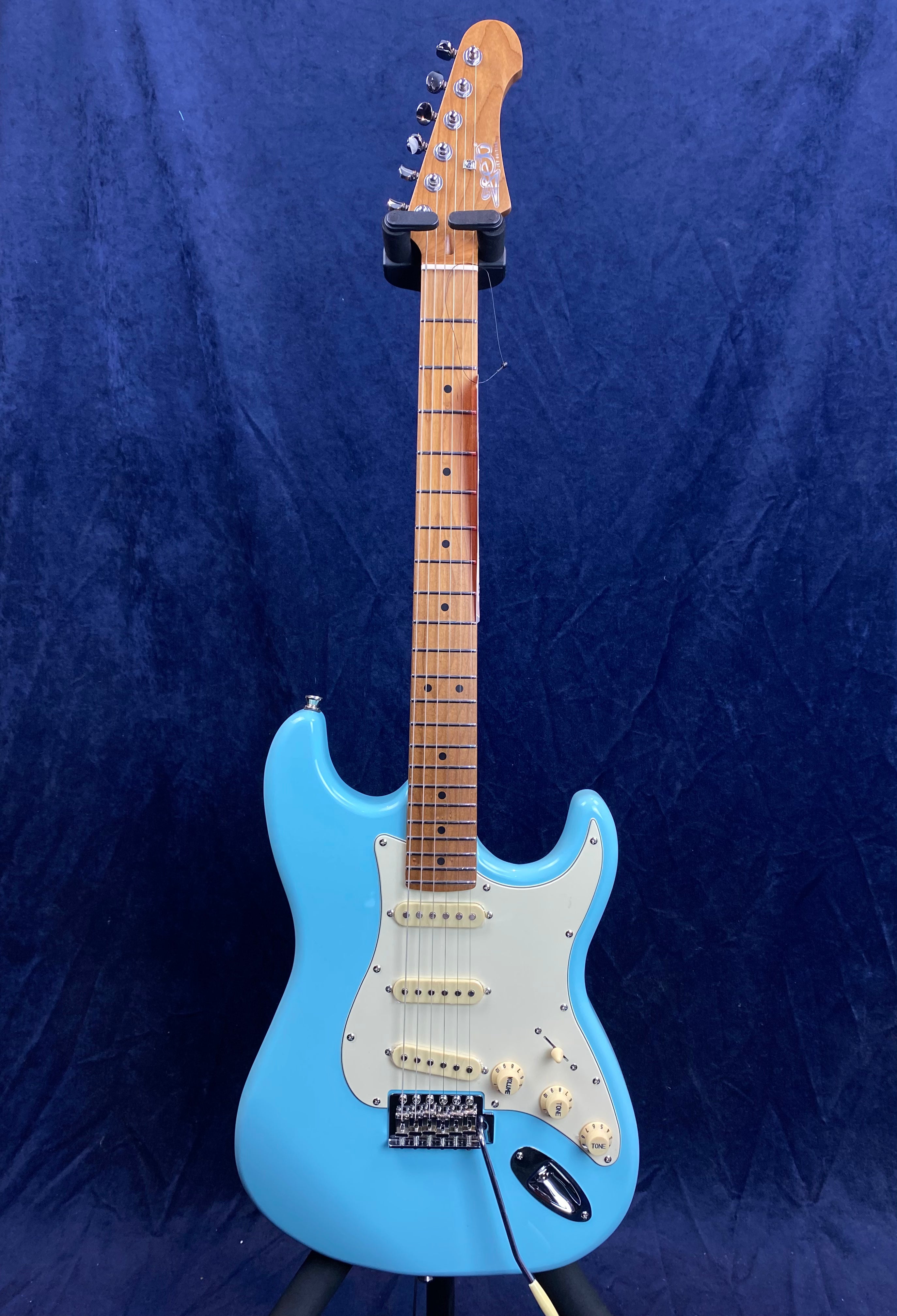 Electric guitar with a light blue body, white pickguard, three single-coil pickups, maple neck, and six tuning pegs, shown upright against a dark background.