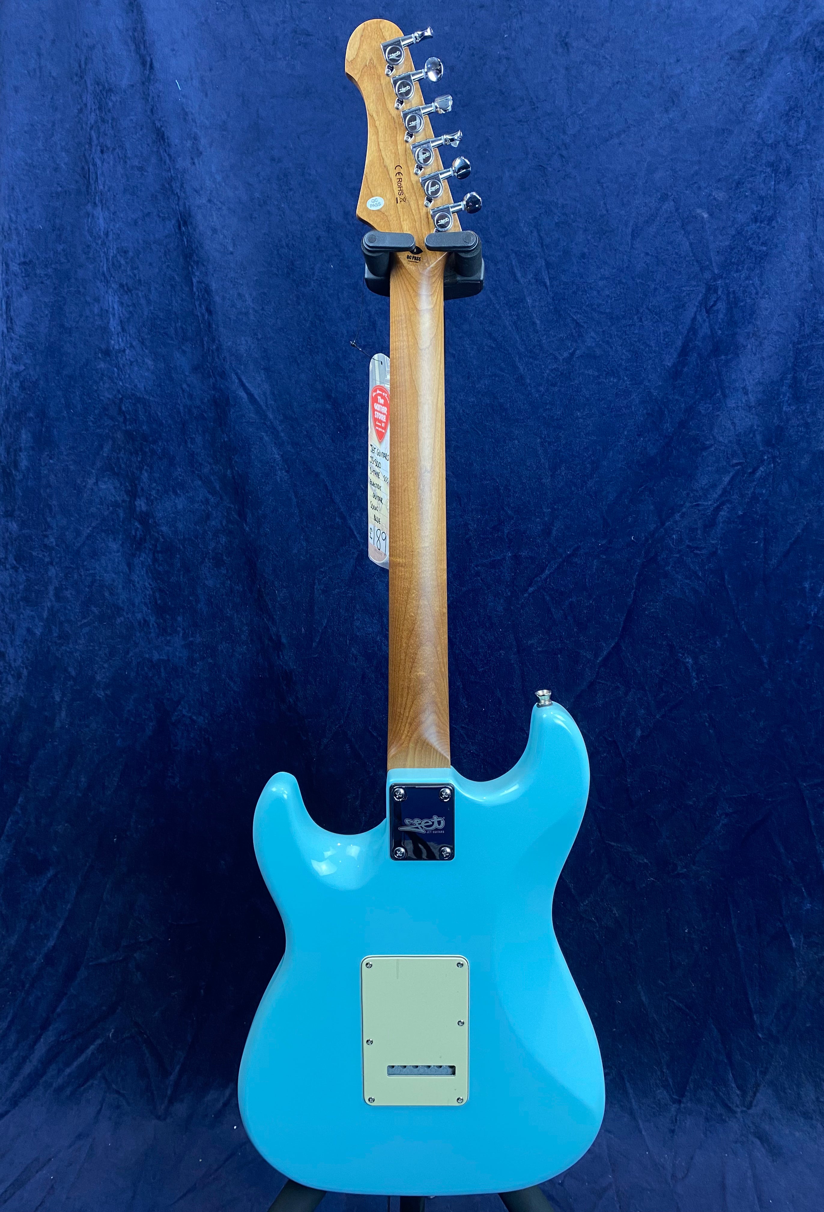 A light blue electric guitar viewed from the back, with a natural wood neck and chrome tuning pegs, set against a dark blue background.