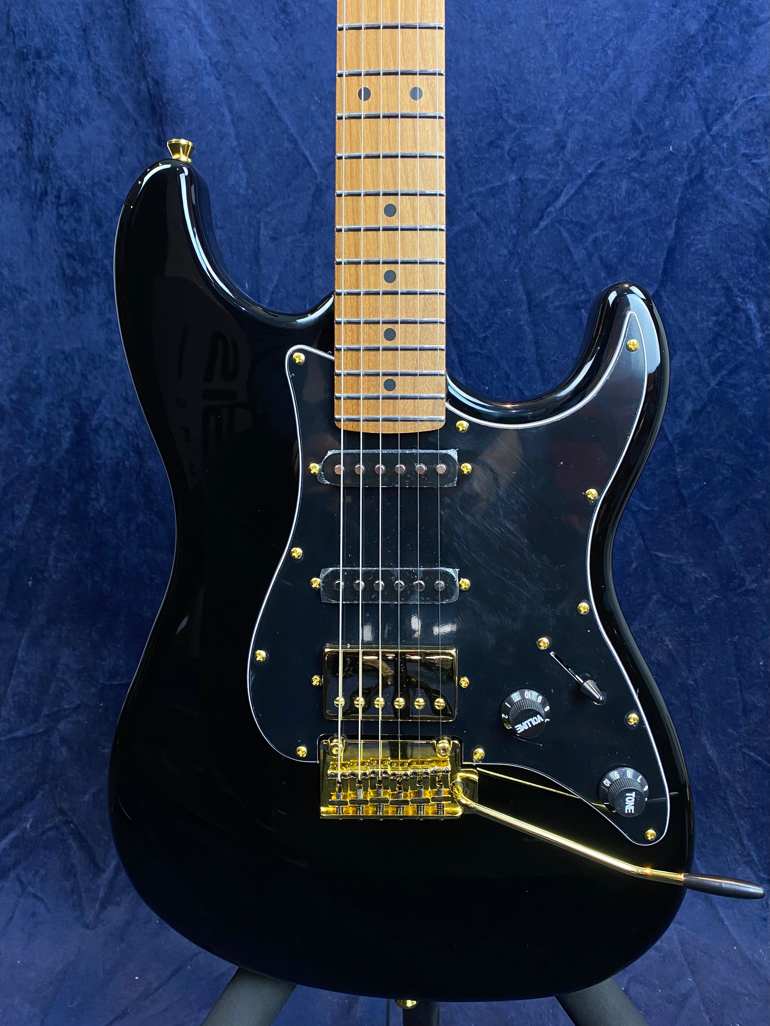 Black electric guitar with gold hardware, three pickups, two black control knobs, and a gold tremolo arm against a dark blue background.