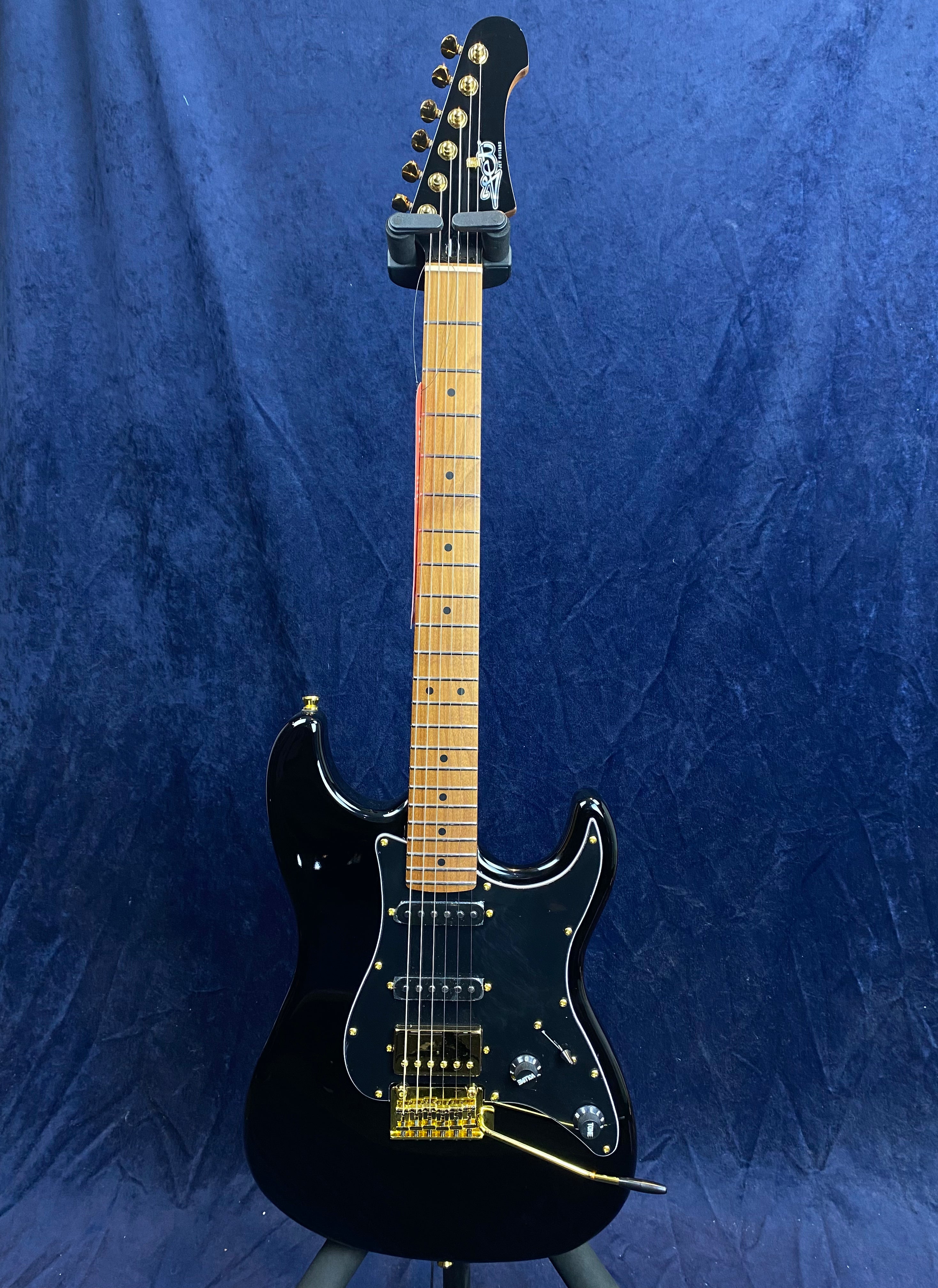 Black electric guitar with maple fretboard, gold hardware, three pickups, two control knobs, and a whammy bar, against a dark blue background.