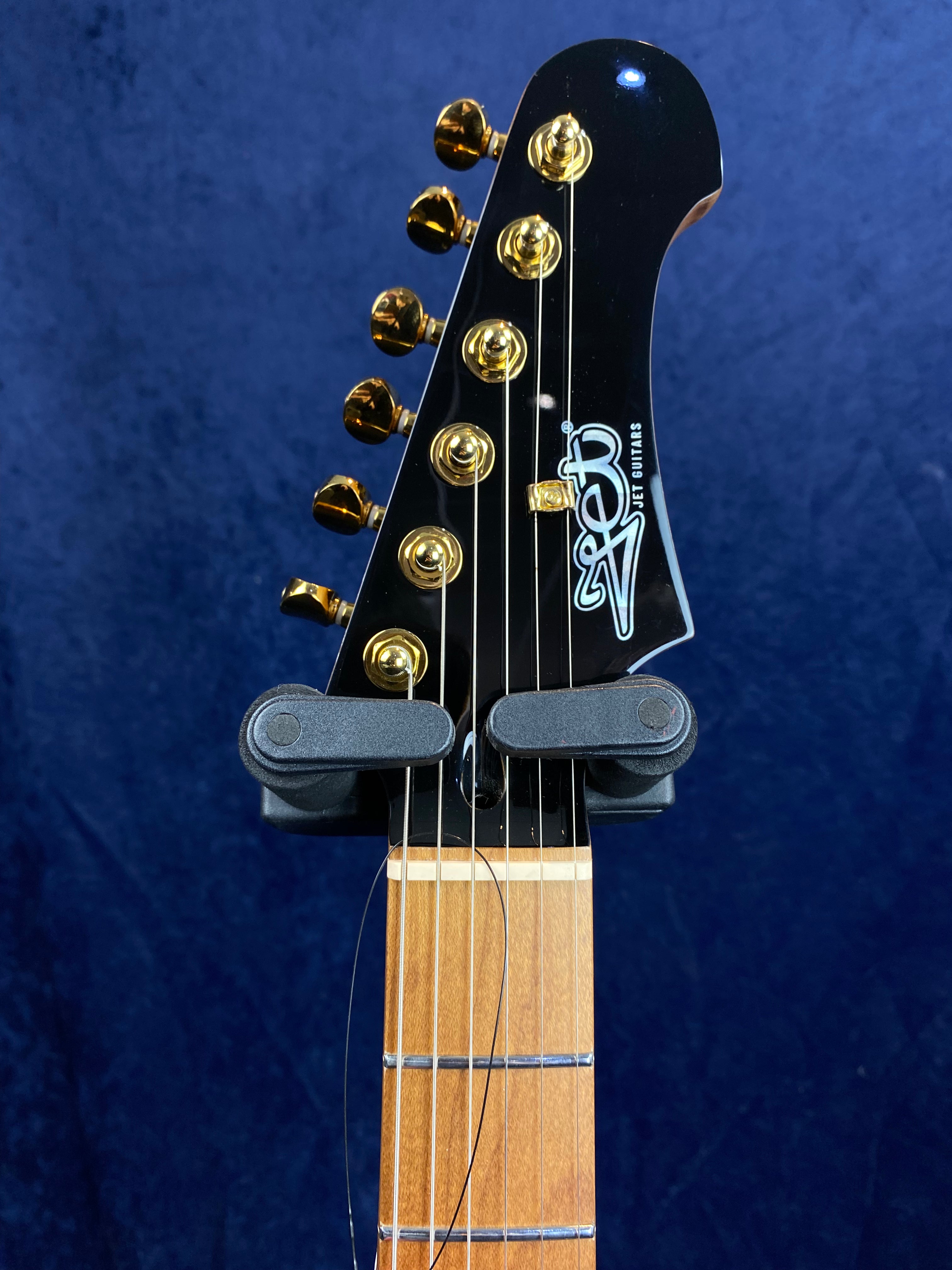 Close-up of an electric guitar headstock with gold tuning pegs, a black finish, and "Jet Guitars" logo, neck secured in a wall hanger.