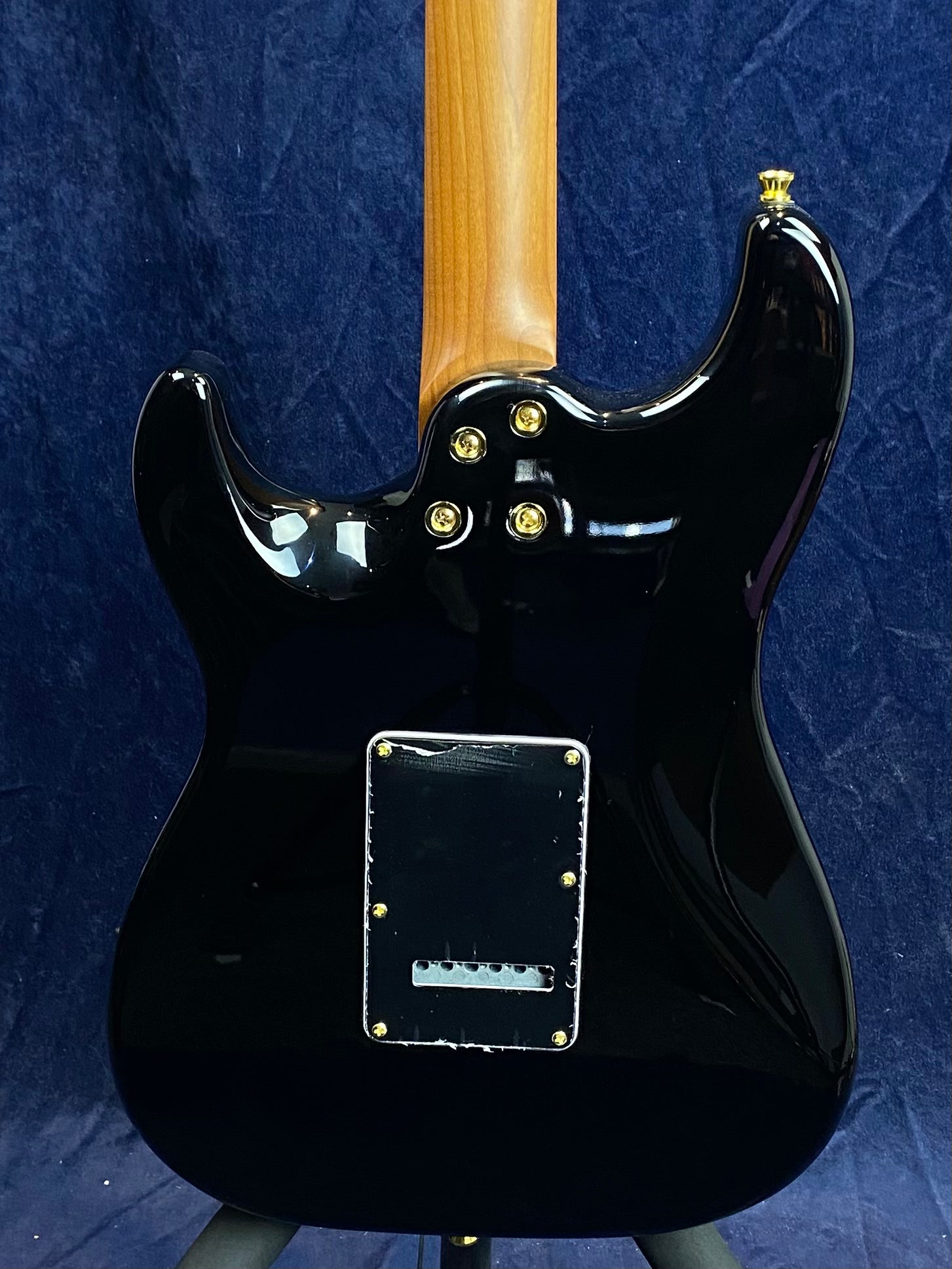 Glossy black electric guitar back with a natural wood neck, gold screws on the neck joint and tremolo cover plate, set against a dark blue background.