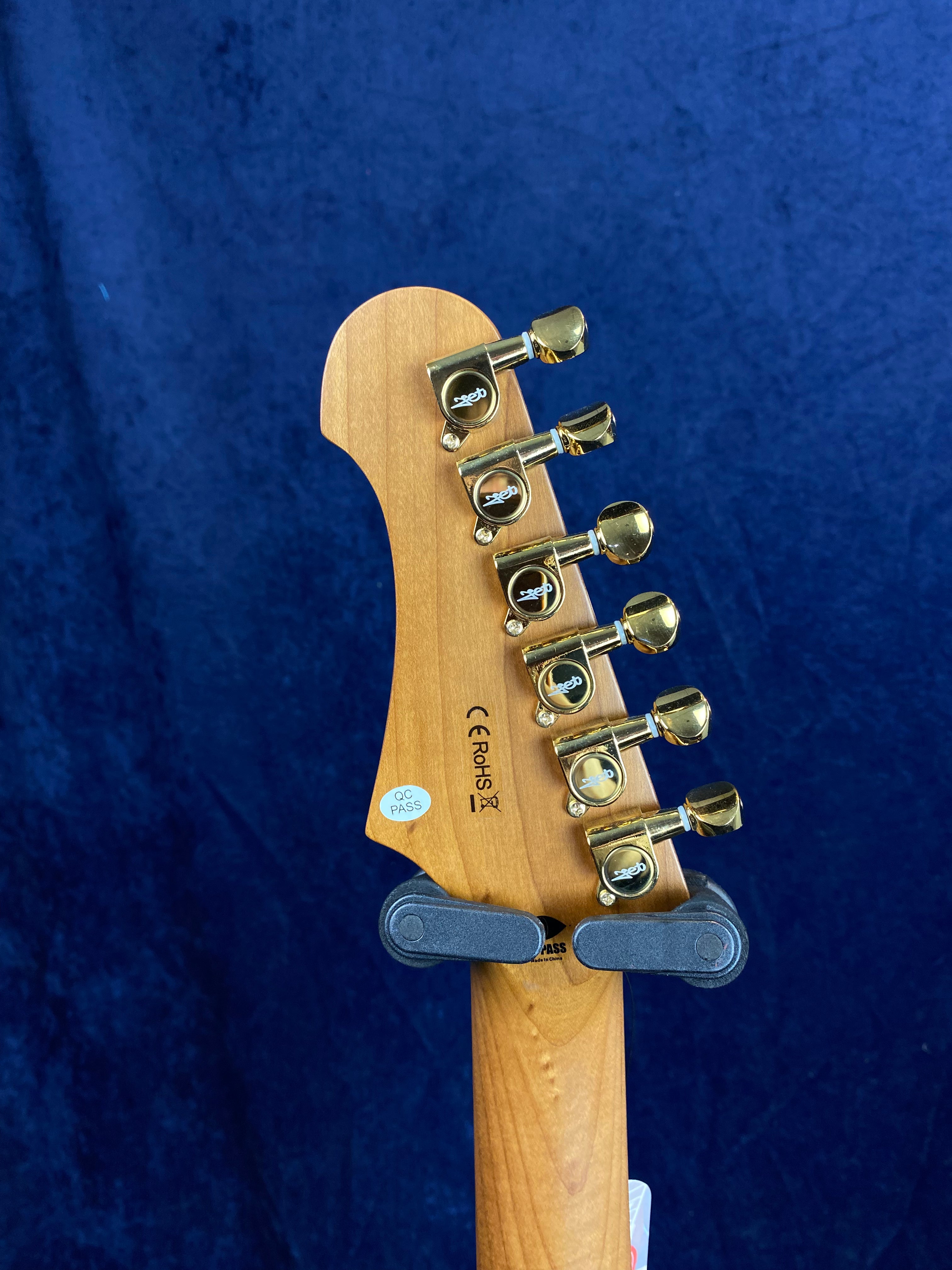 Back of an electric guitar headstock showing six gold tuning pegs, QC and certification stickers, against a dark blue background.