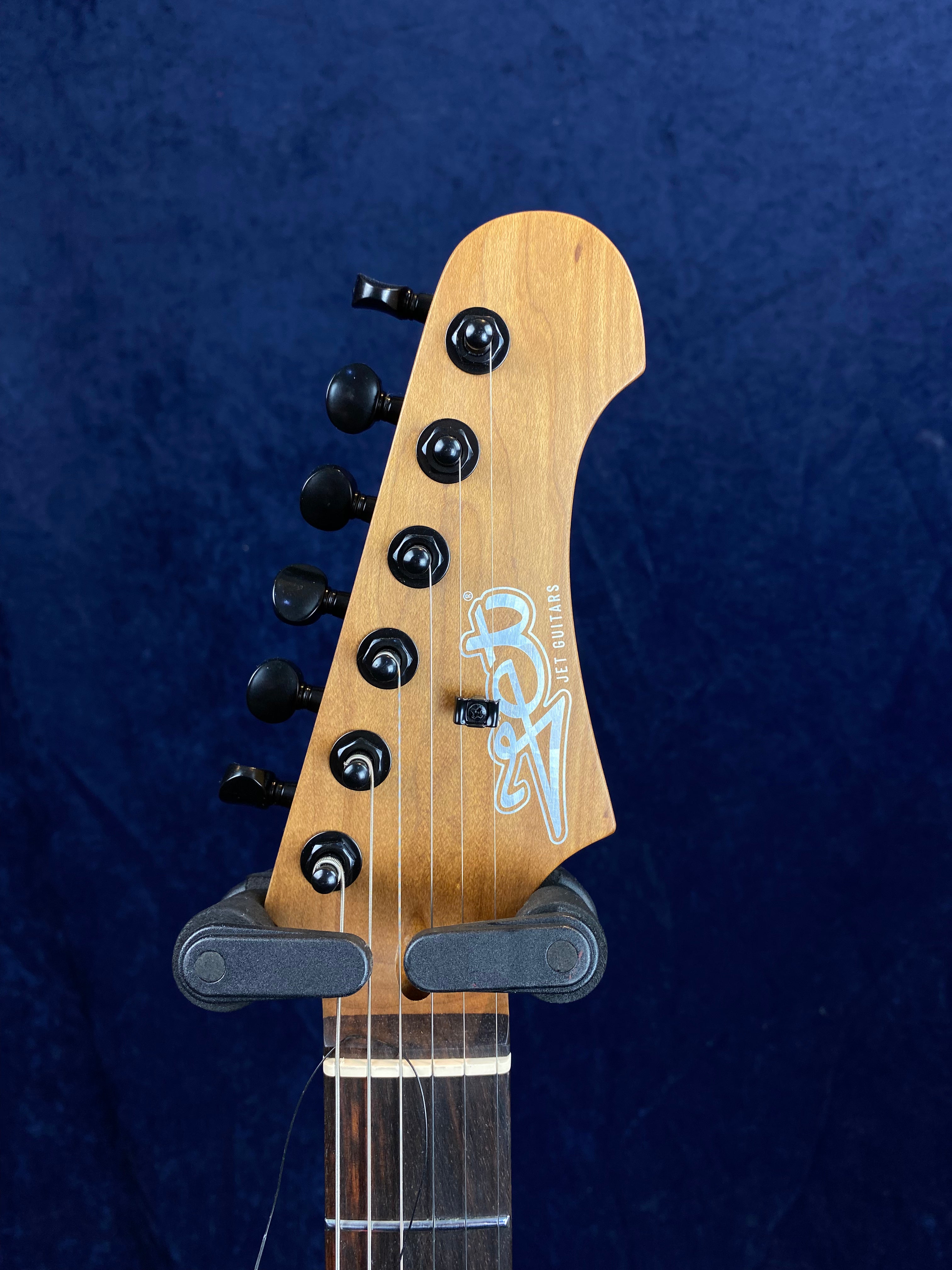 Light brown electric guitar headstock with six black tuning pegs and a stylized "Jet Guitars" logo on the front, shown against a dark blue background.