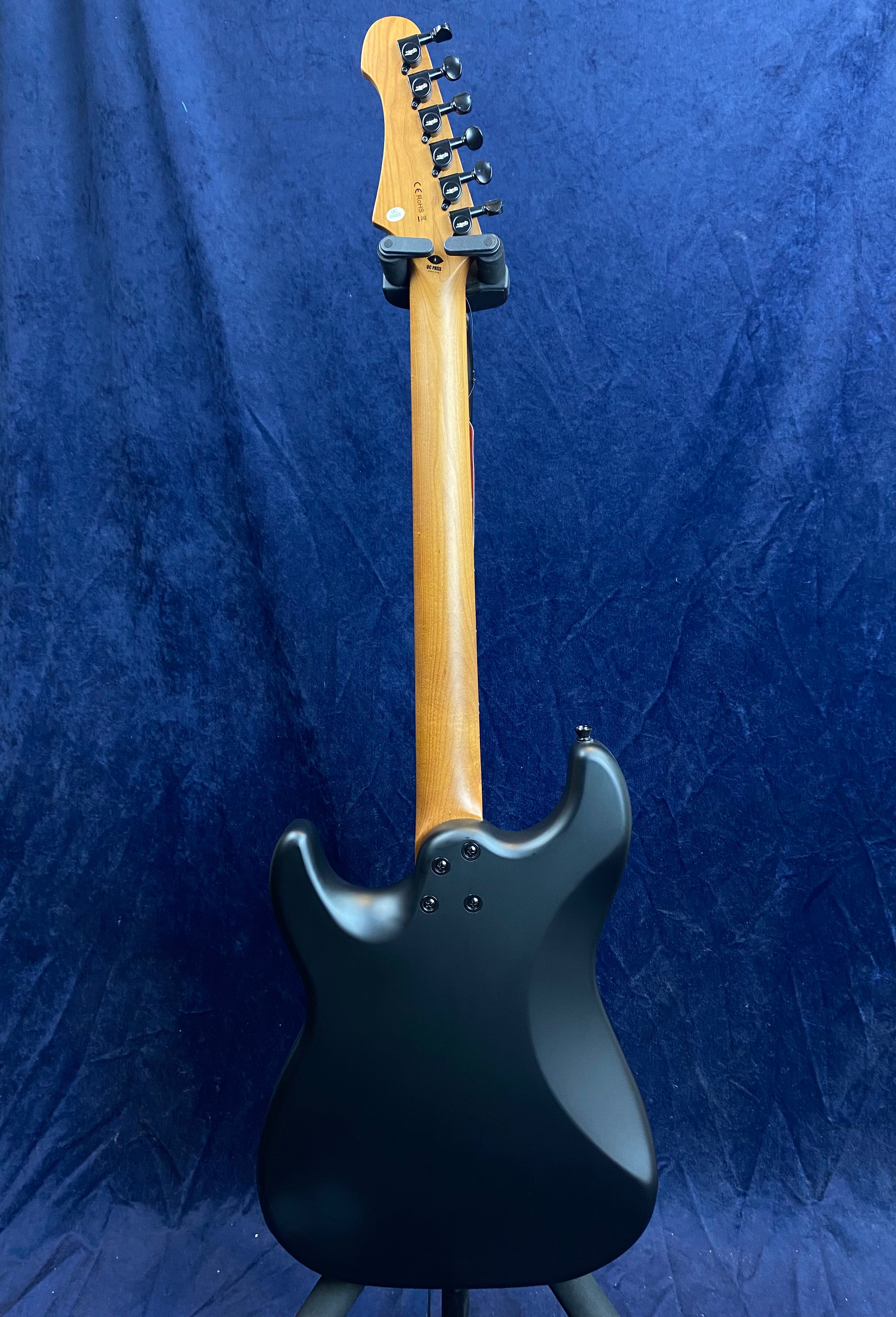 Black electric guitar shown from the back, with a natural wood neck and six tuning pegs on the headstock, set against a dark blue background.